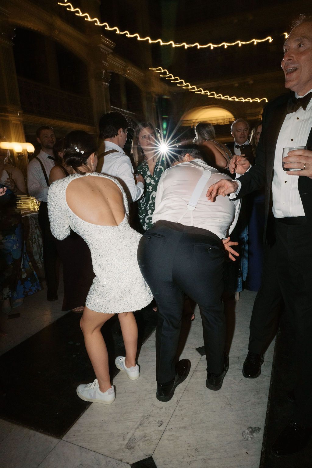 Wedding party members dancing in formal attire under decorative string lights.