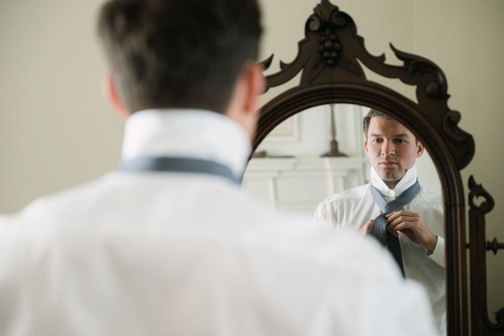 Groom adjusts tie while looking in an ornate dark wooden mirror.
