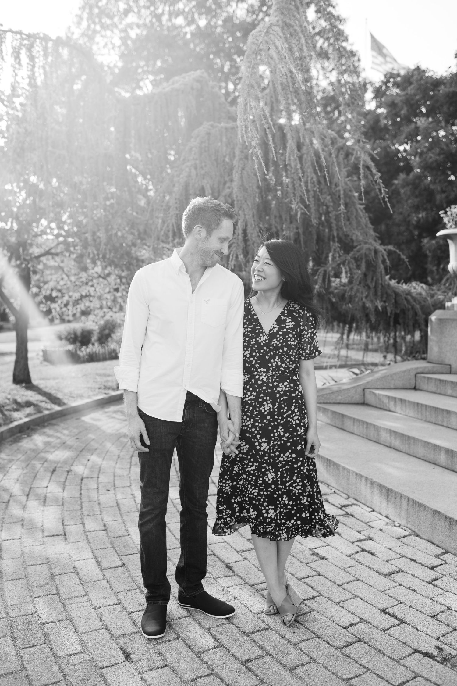 Black and white photo of a smiling couple walking on a brick path surrounded by lush trees and stone steps.