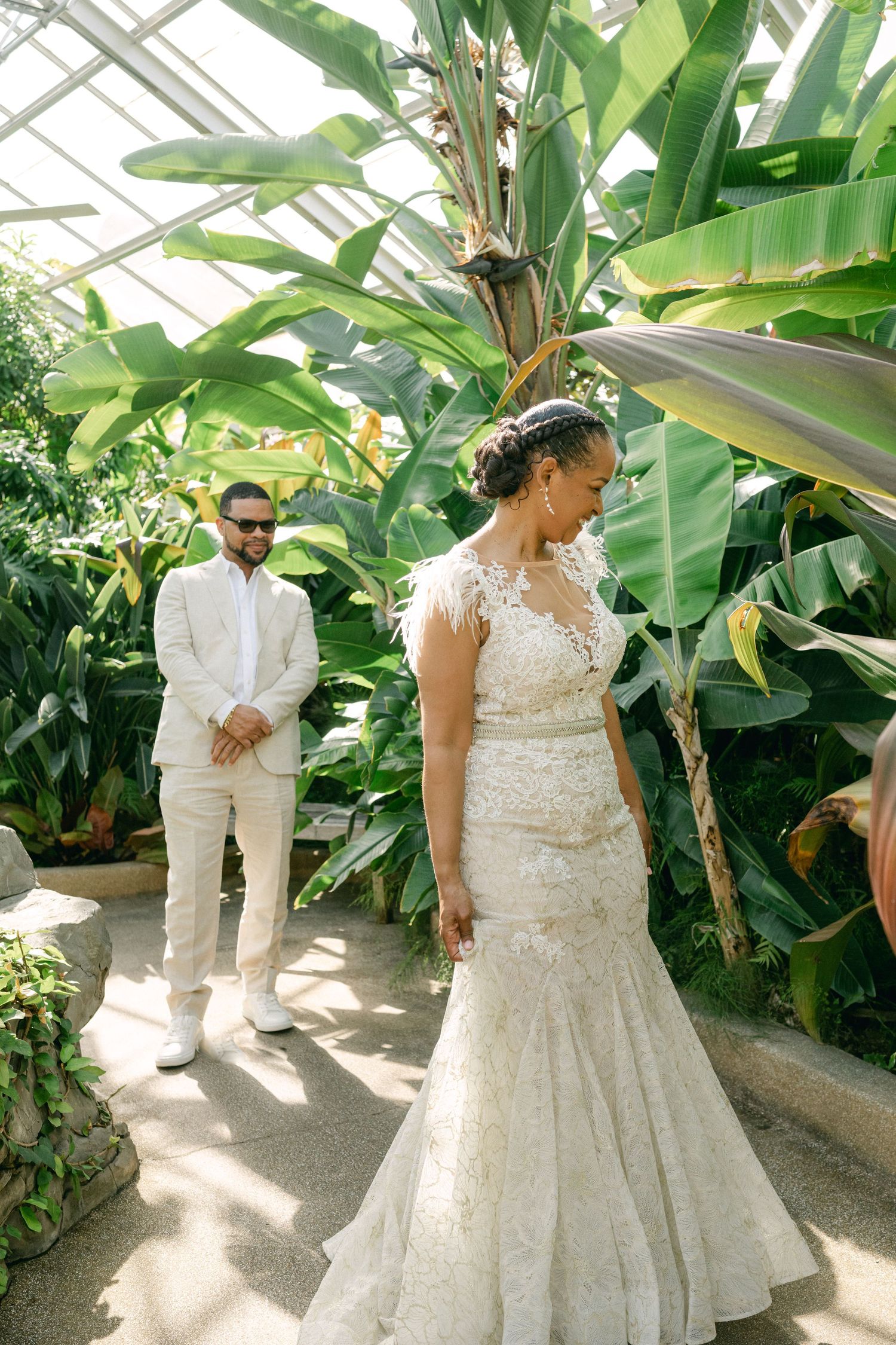 Bride in lace mermaid gown stands before groom in white suit amid lush tropical plants in a sunlit greenhouse.