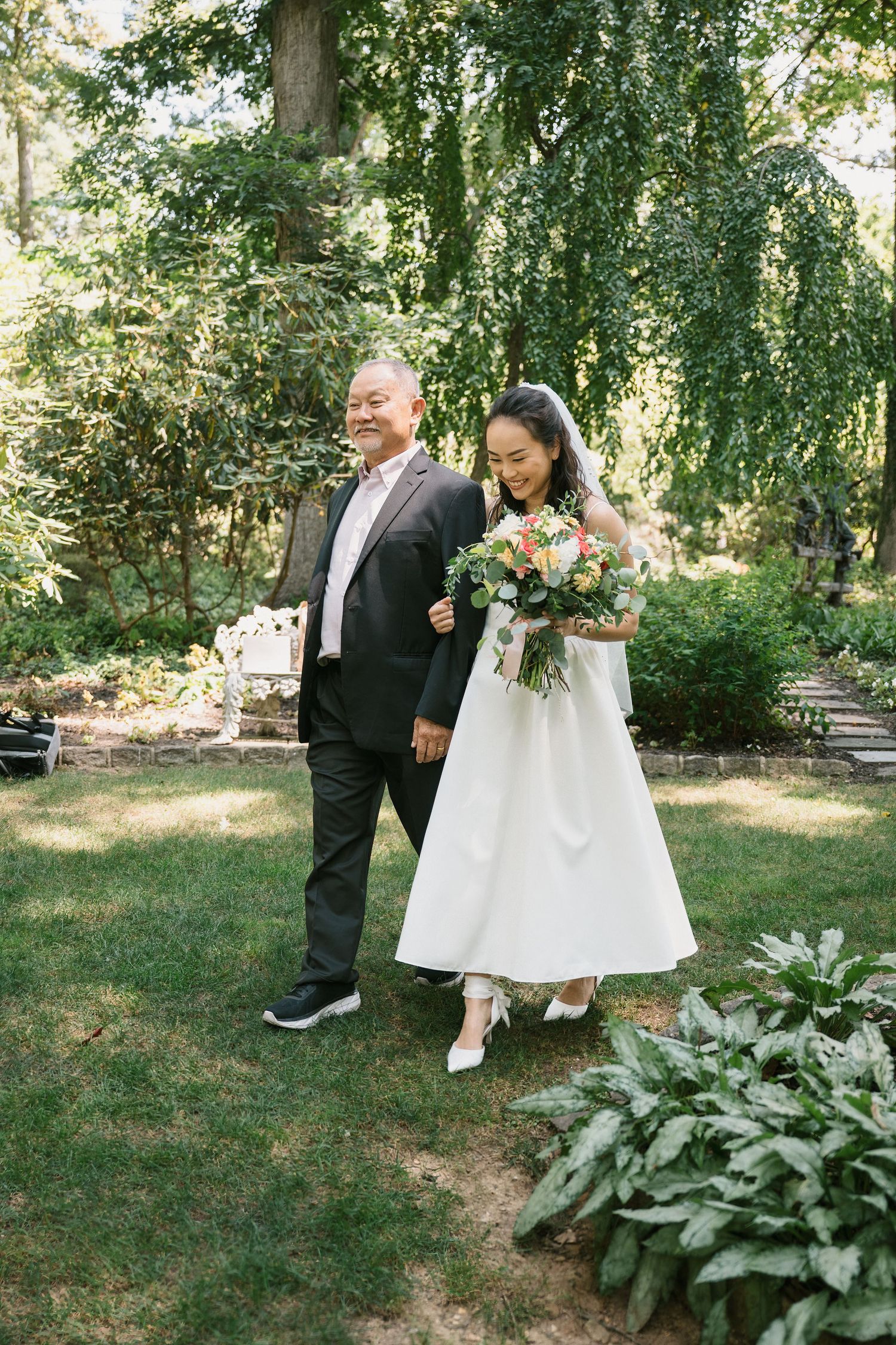 Wedding party walks through a serene garden setting on a sunny day.