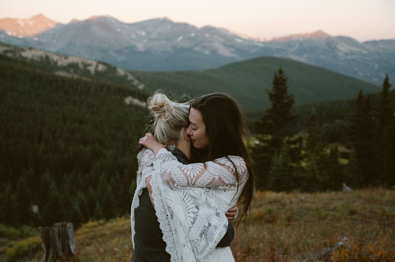 Two women sharing a romantic kiss at sunset with mountain and pine tree backdrop.