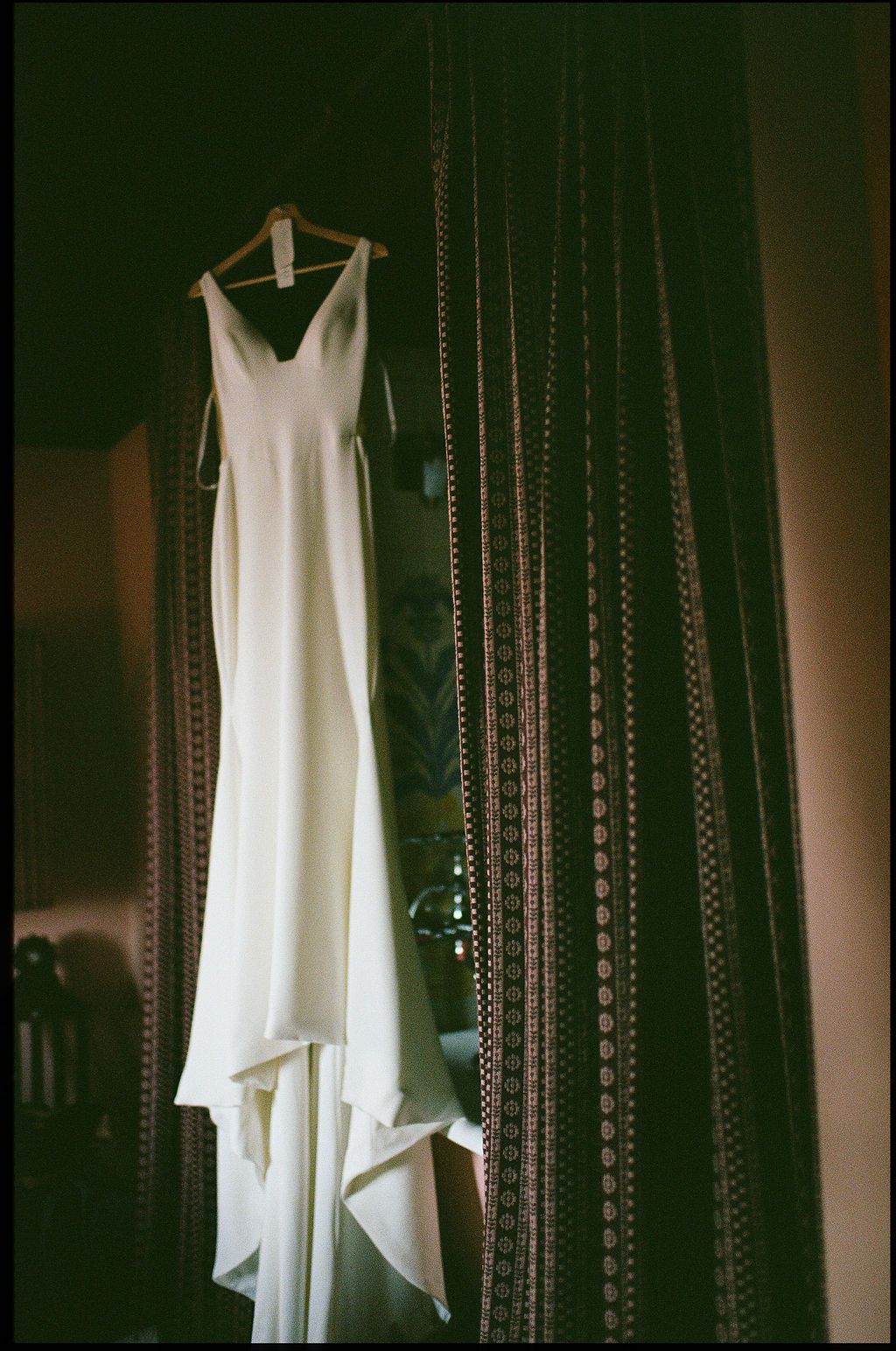 A white wedding dress hangs elegantly against dark patterned curtains in a dimly lit room.