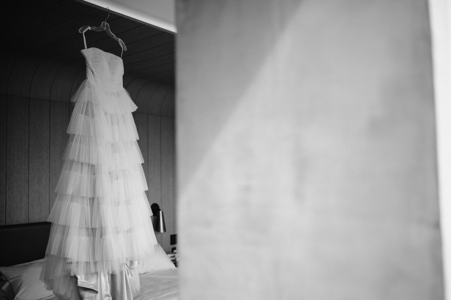 A layered white wedding dress hangs elegantly against a wall in black and white.