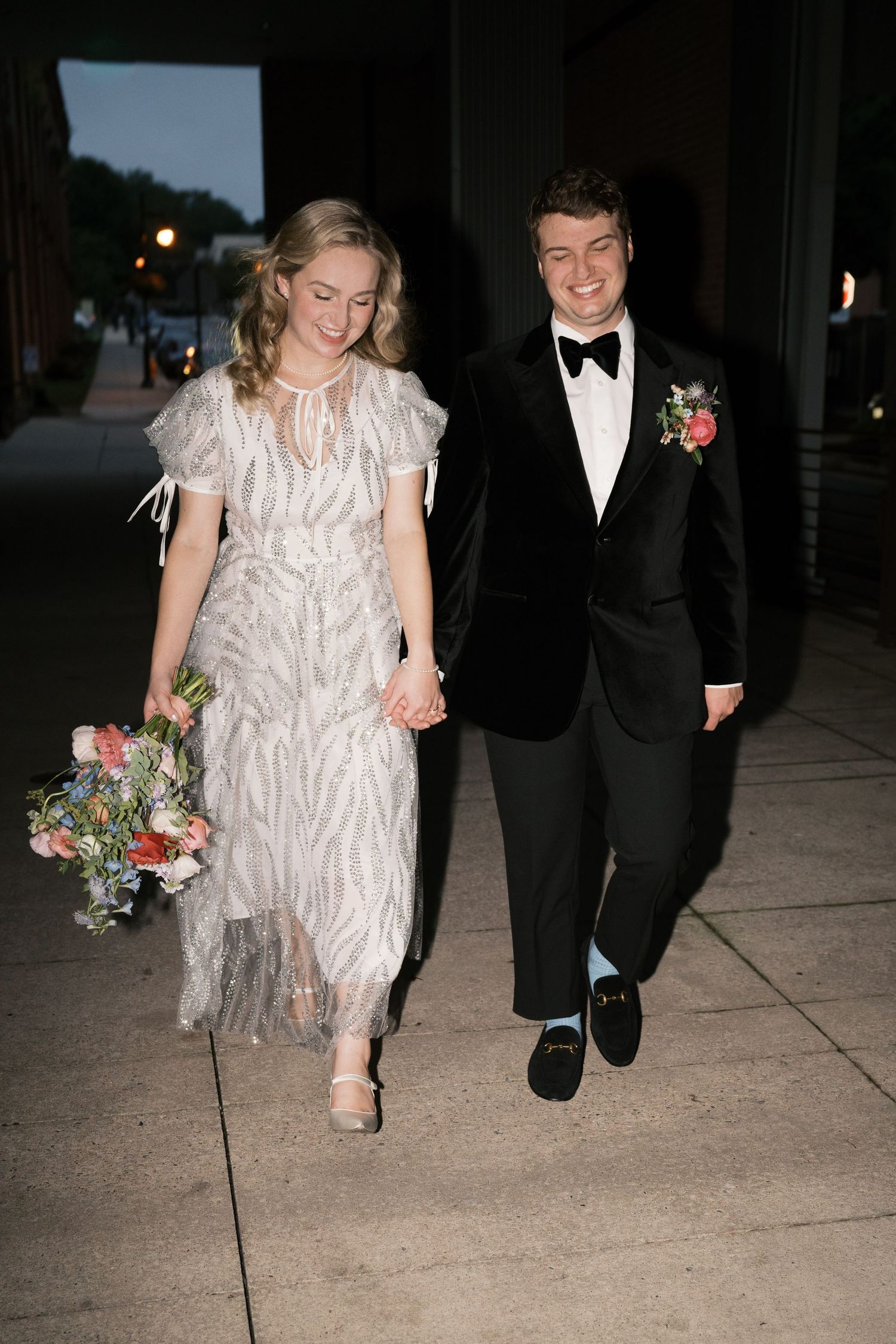 A couple walking hand in hand at night, bride in vintage-style dress and groom in classic tuxedo.
