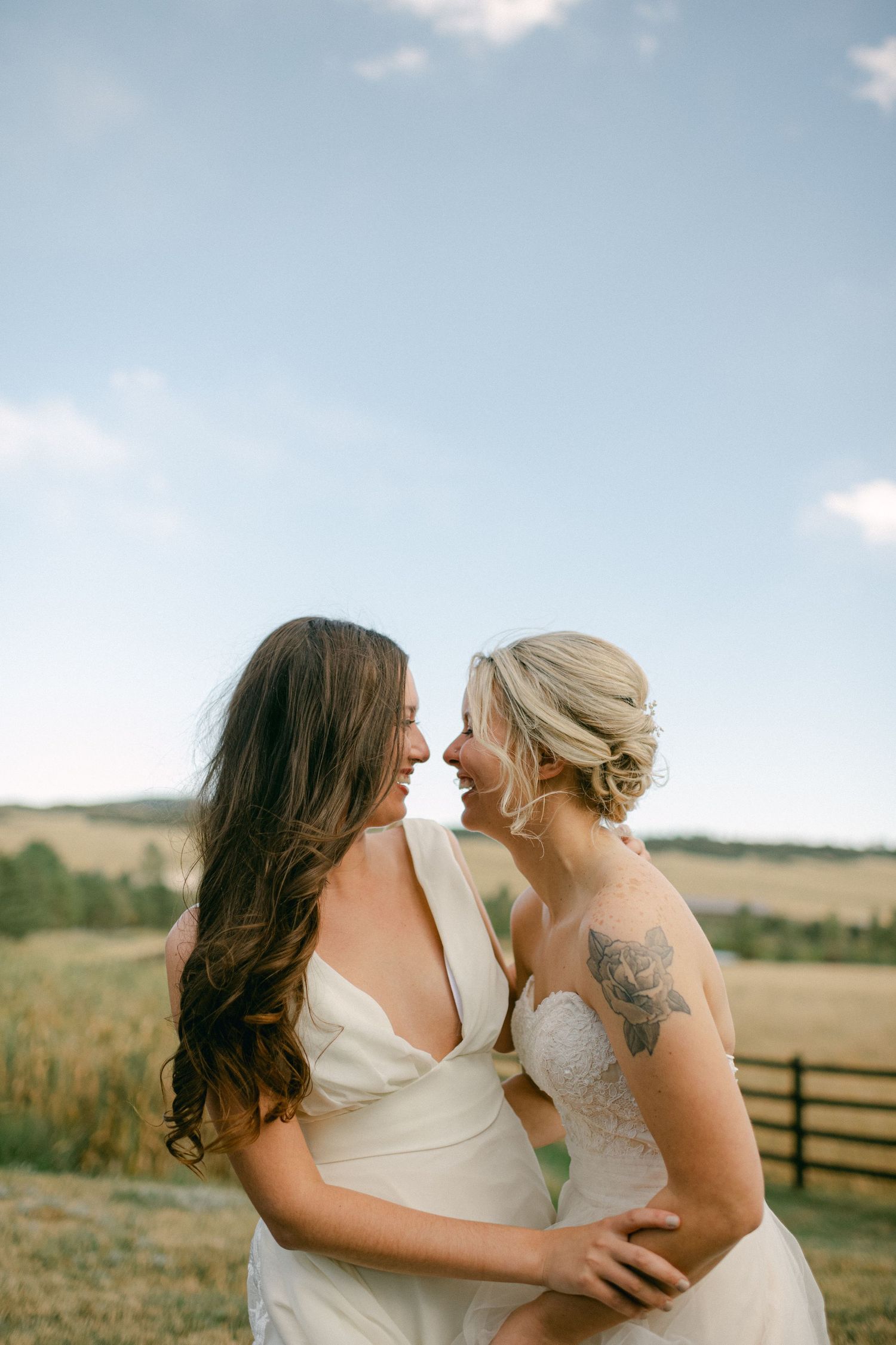 Two people in white dresses share an intimate moment against a rural backdrop with rolling hills and fence.