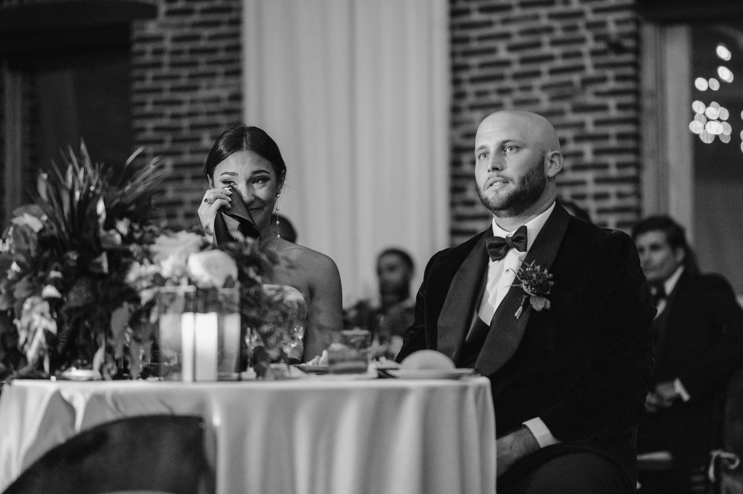 A couple sits at their reception table with candlelight and floral arrangements during an elegant indoor wedding celebration.