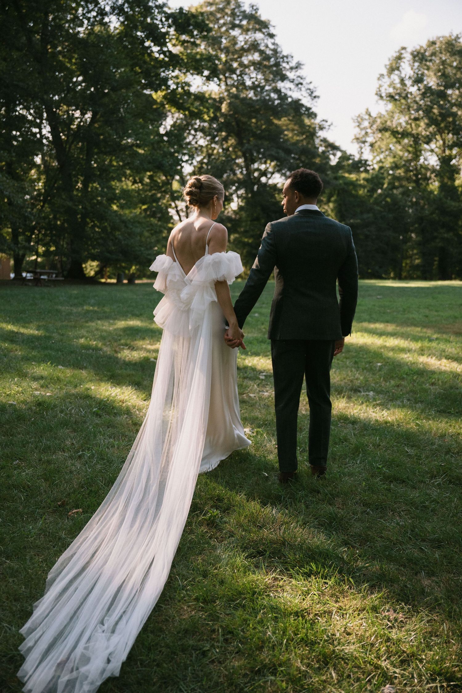A wedding couple stands in a sunlit garden as the bride's long flowing veil trails behind them.