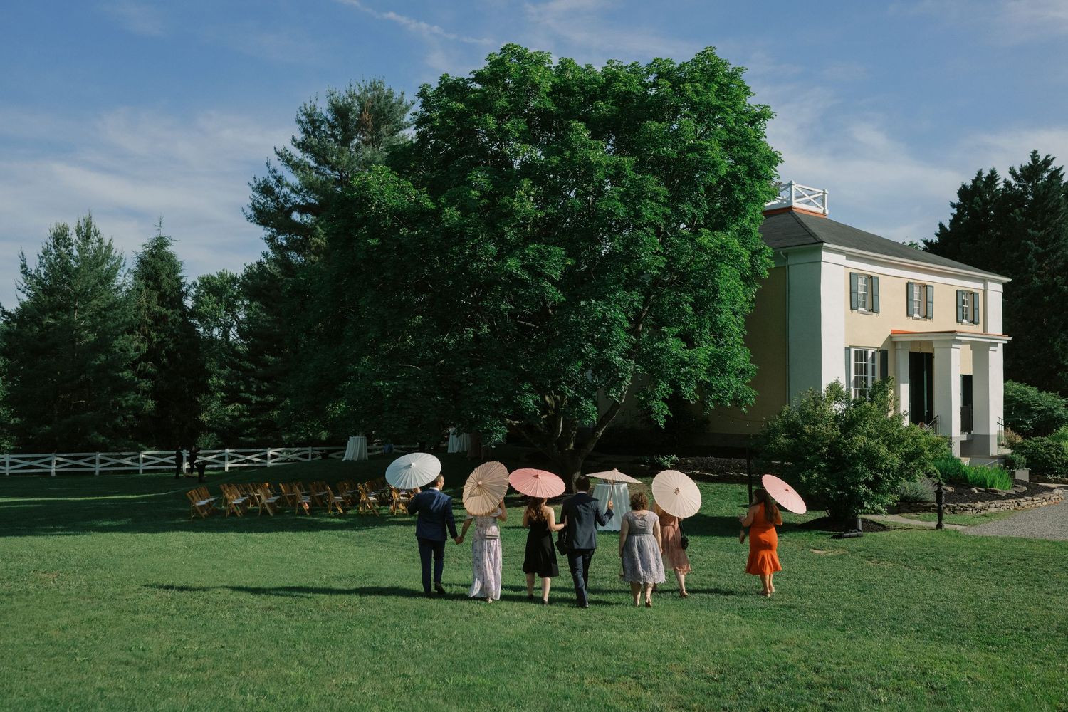 A group with parasols stands on a lawn in front of a white colonial-style house on a sunny summer day.