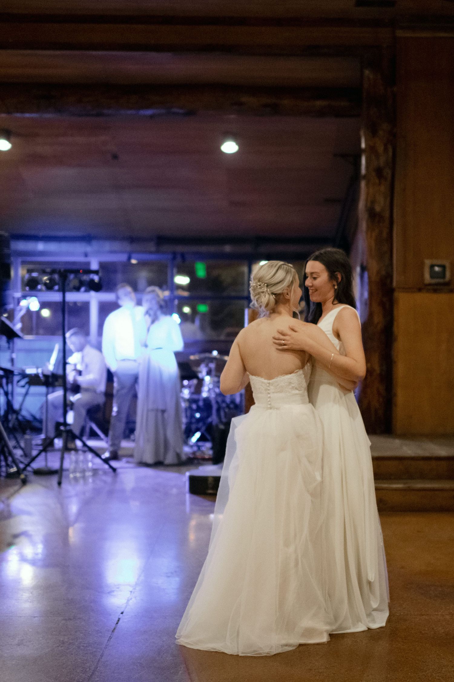 Two people in elegant white dresses share an intimate dance moment at a wedding reception with a band in the background.