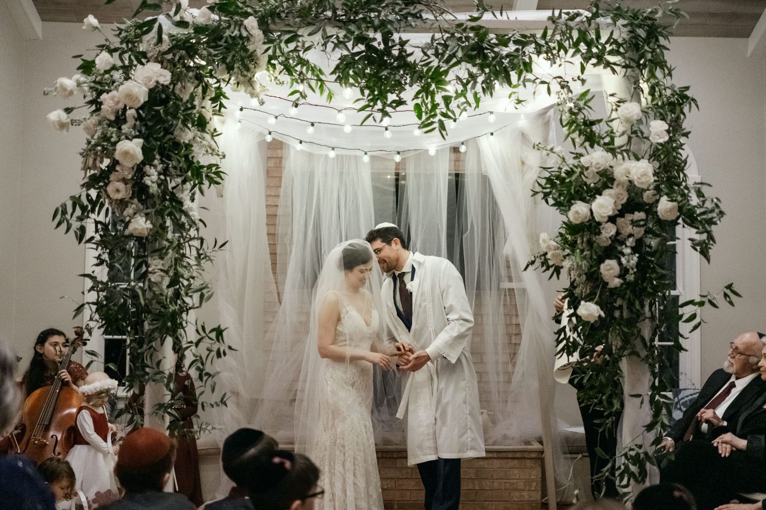Intimate indoor wedding ceremony with floral arch, string lights and guests seated in viewing area.