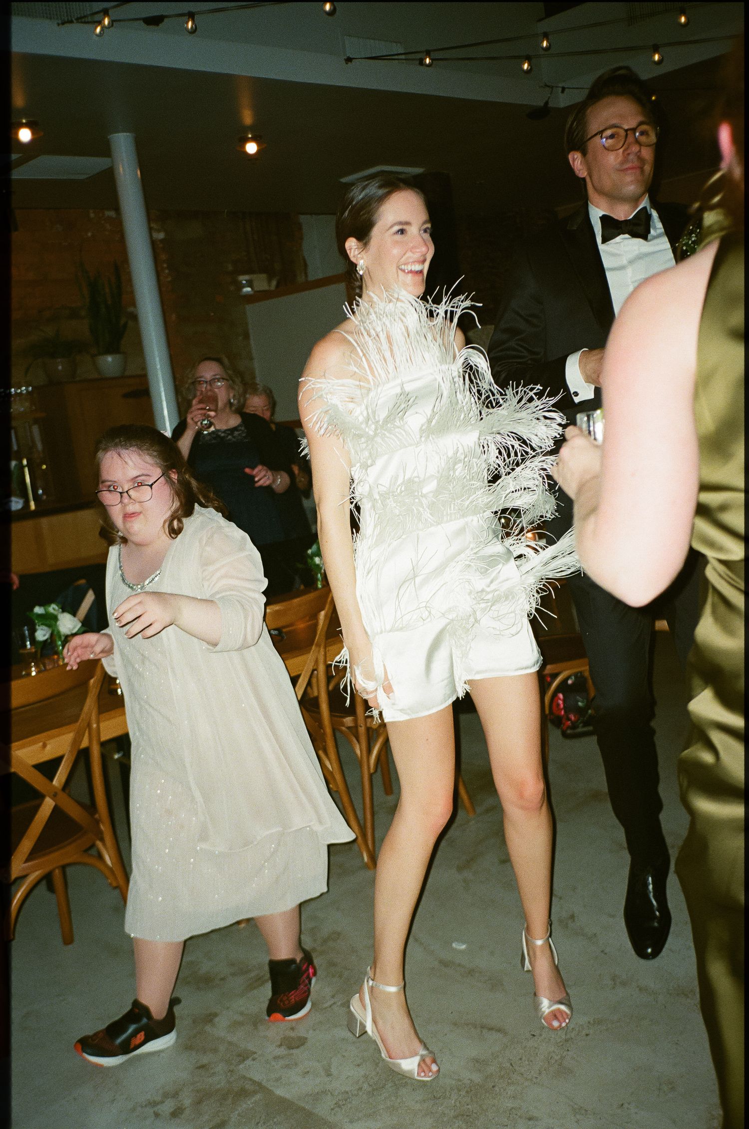 Party guests dance and celebrate at an event with people wearing formal attire including a white feathered dress.