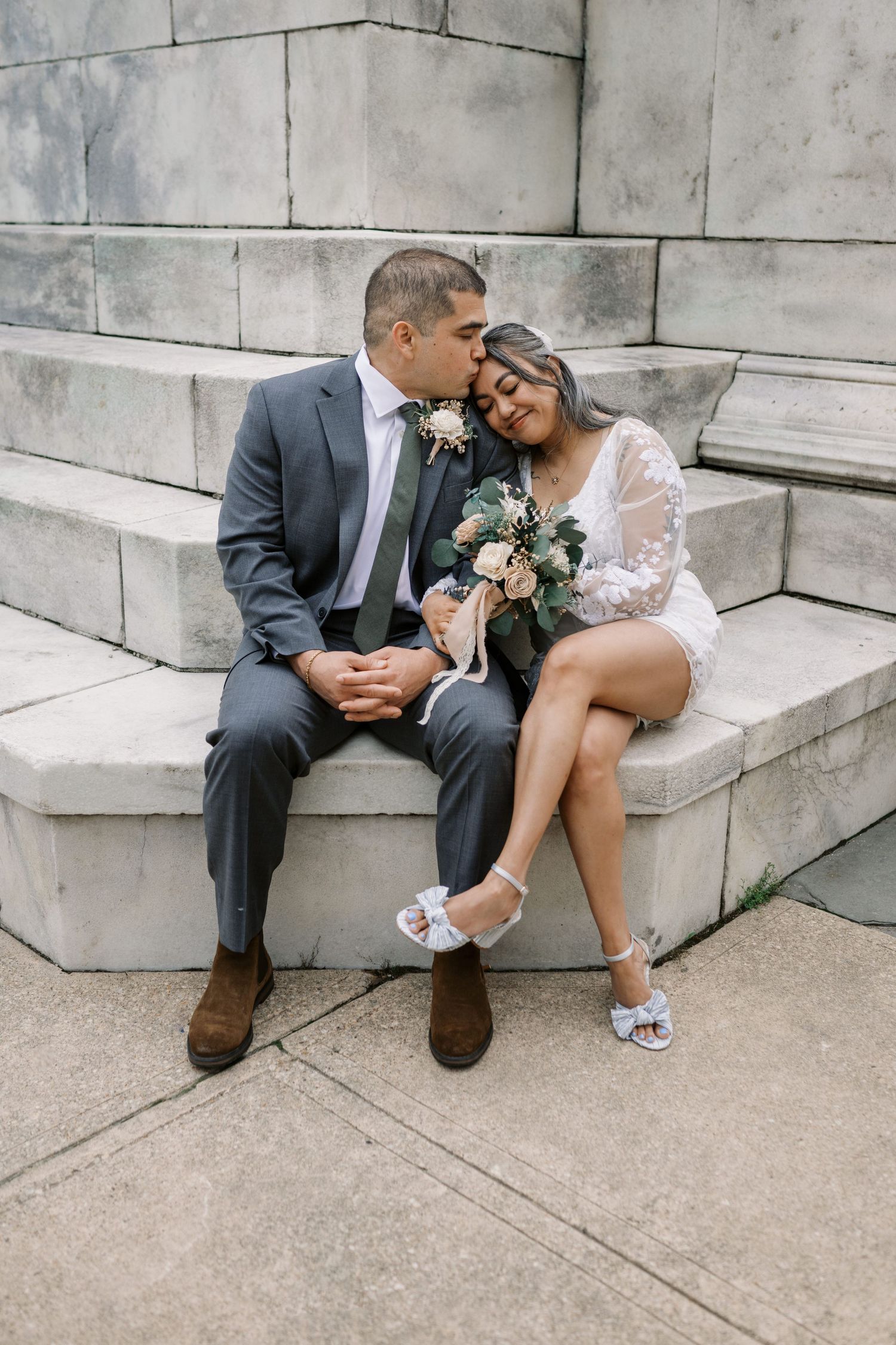 A romantic moment captured between a couple sitting on stone steps in formal wedding attire.