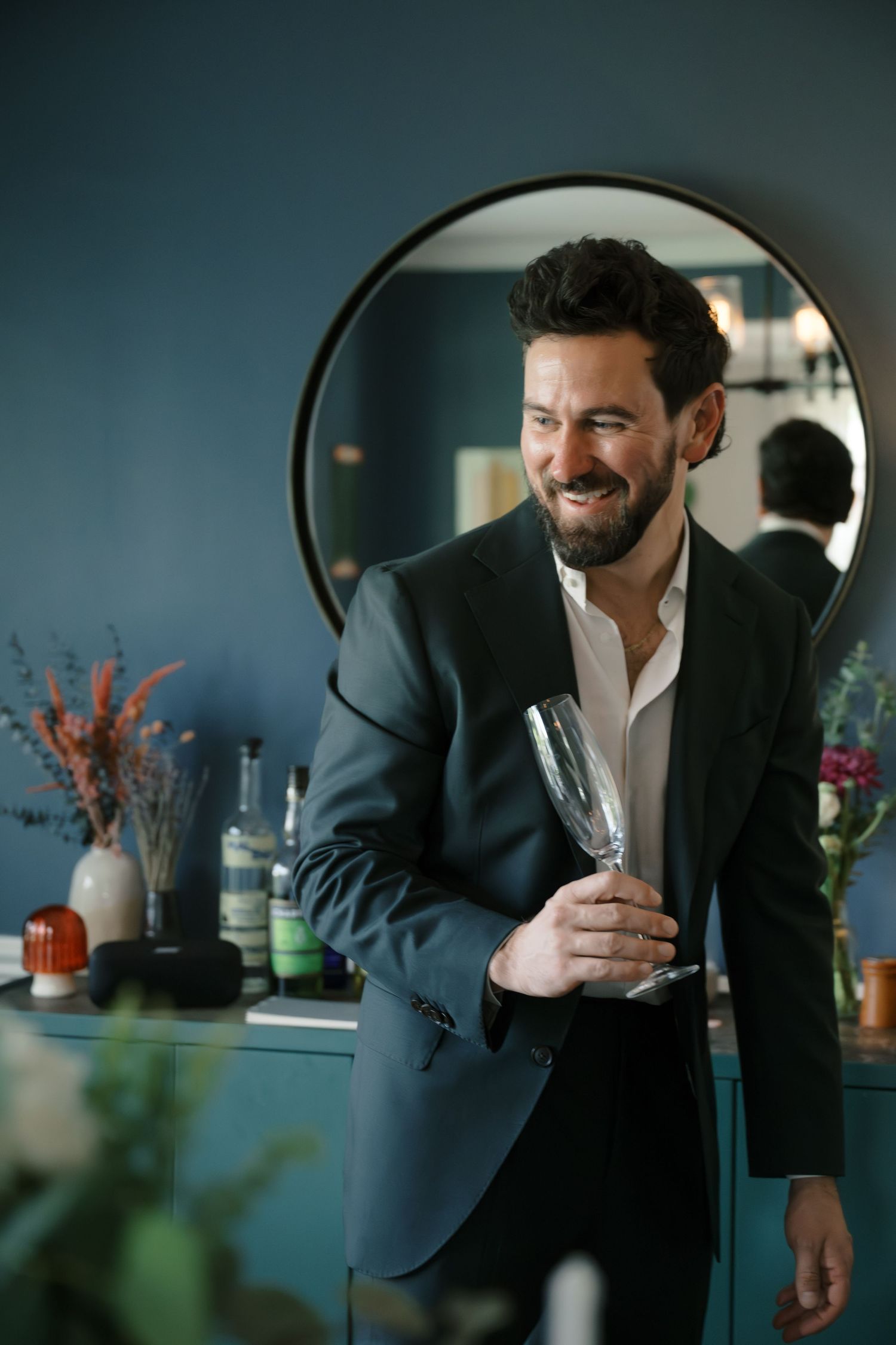 Man in dark suit sips from a glass while standing in a modern room with floral arrangements.