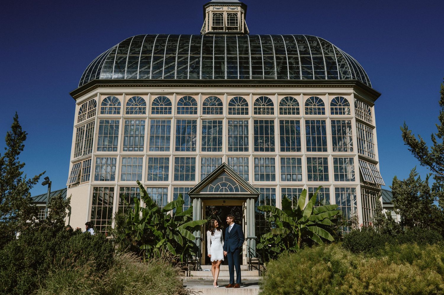 A classic glass conservatory building with white trim and arched windows surrounded by garden landscaping.