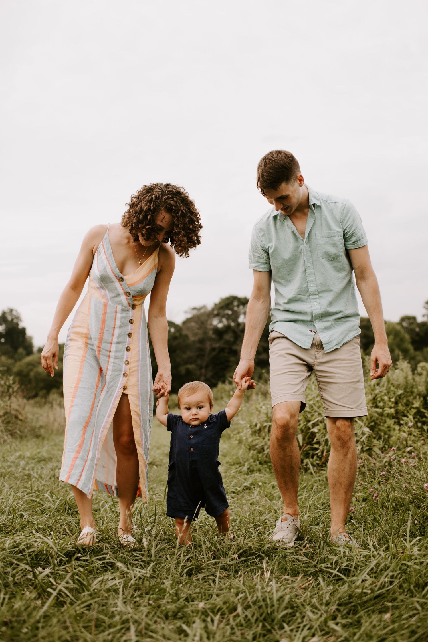 Family helping baby take first steps in grassy field during summer.
