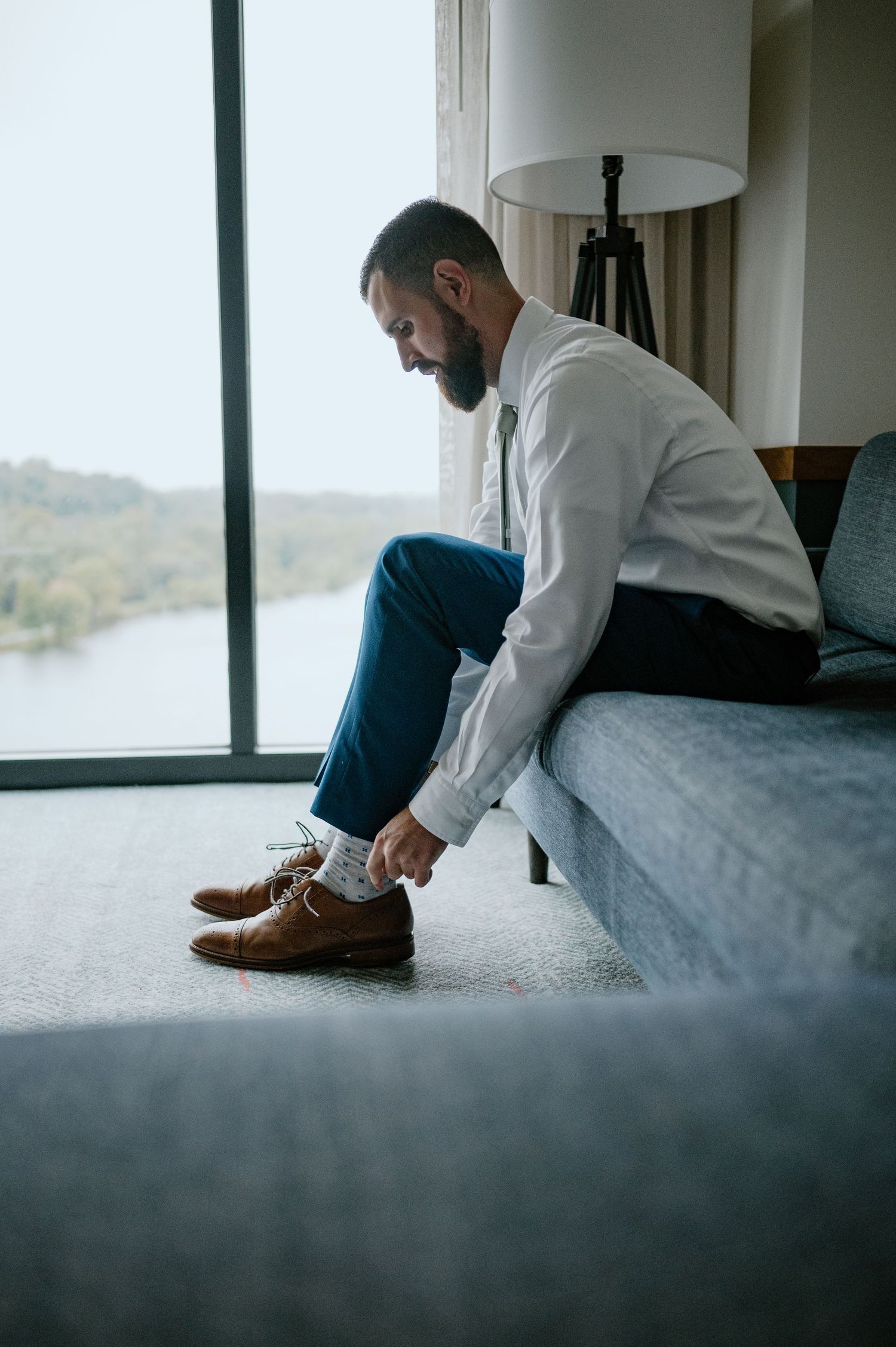 Person in white shirt and jeans tying brown leather shoes while sitting on grey couch near window.