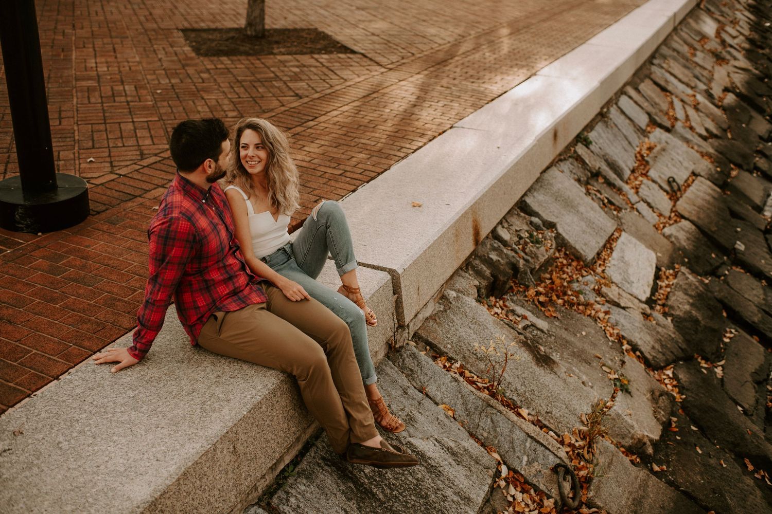 Couple sitting on stone wall by waterfront in casual autumn attire.