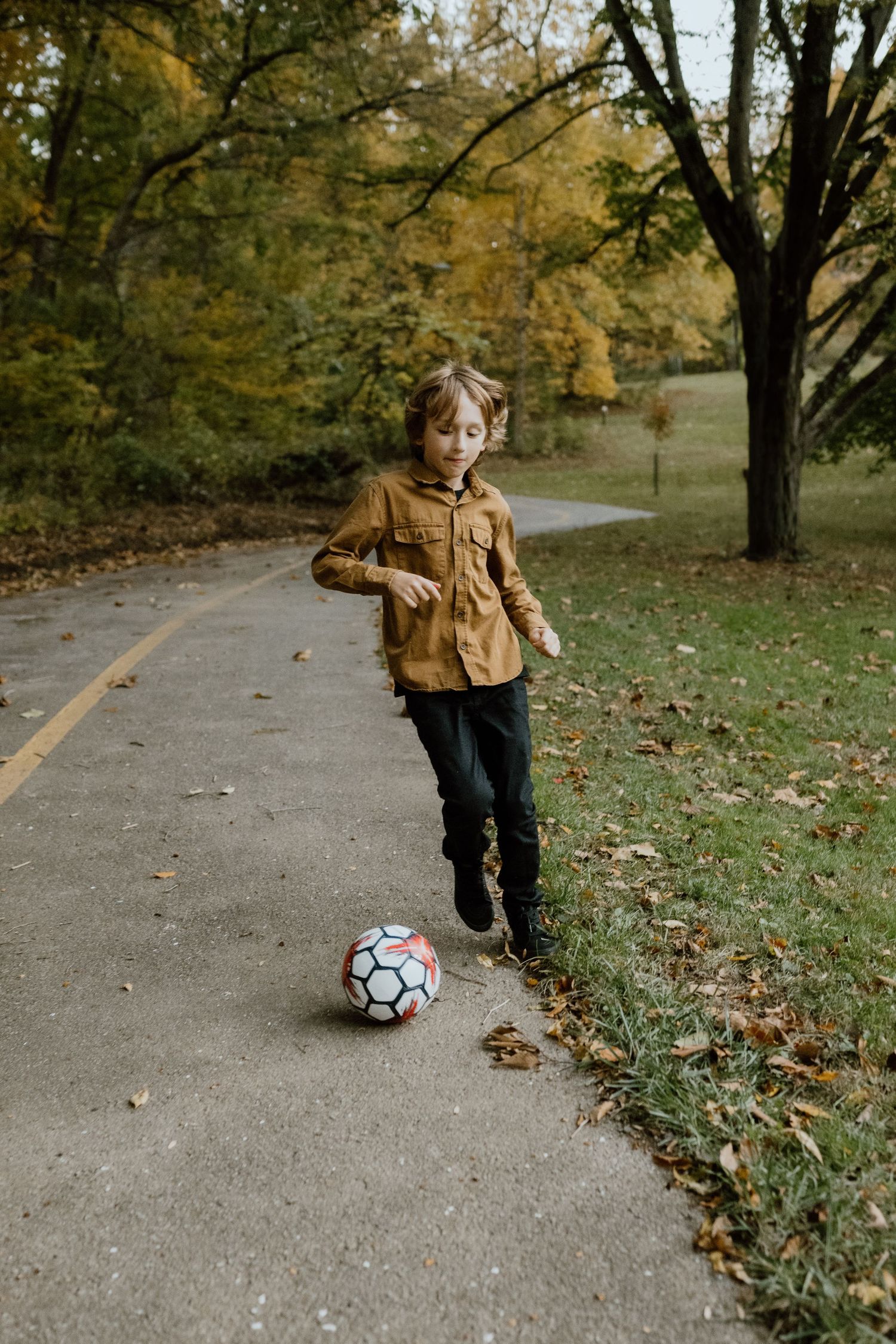 Child in brown jacket plays with soccer ball on path surrounded by fall foliage.