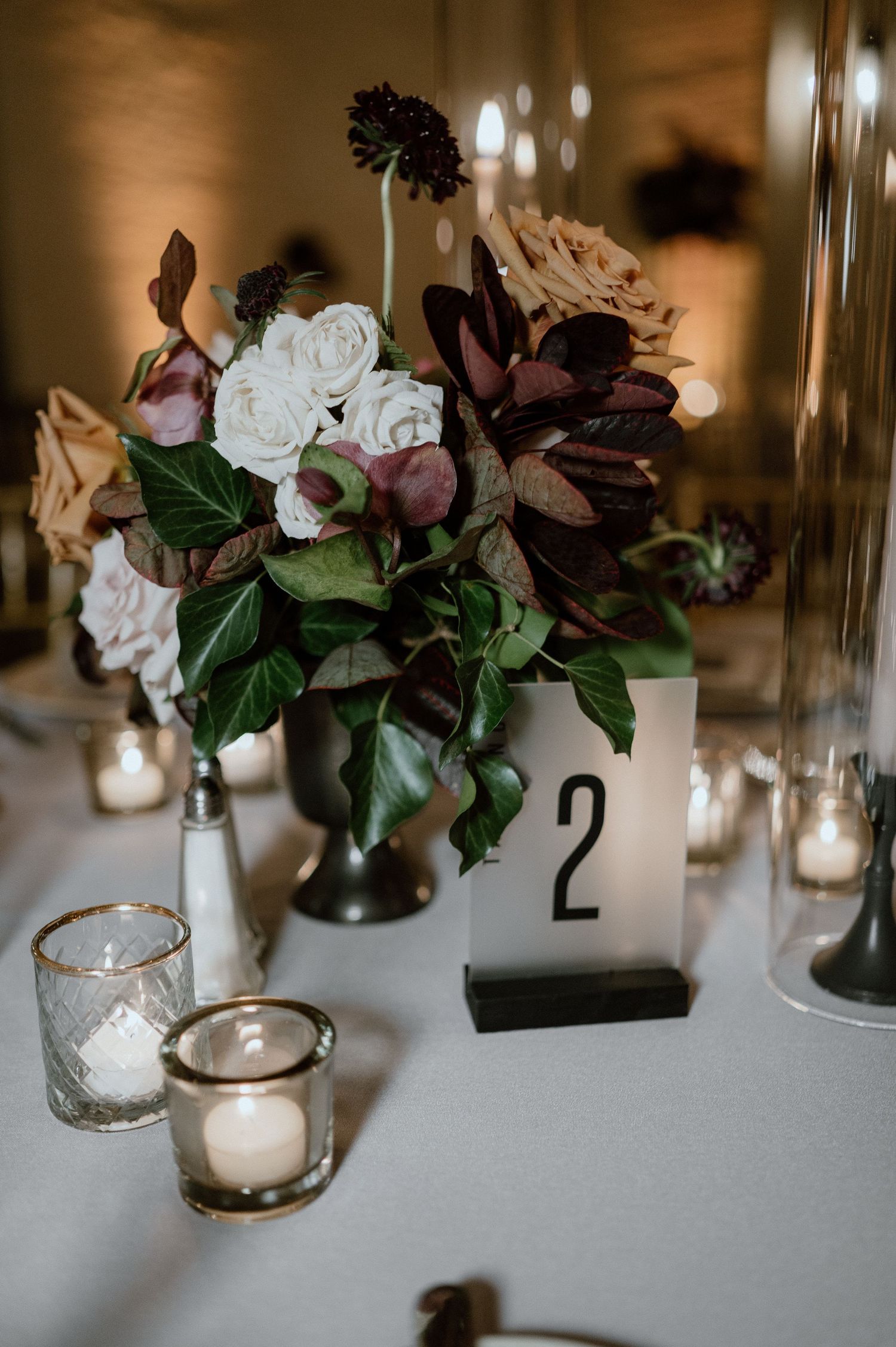 Moody wedding centerpiece with dark burgundy and white flowers alongside table number two and glowing votives.