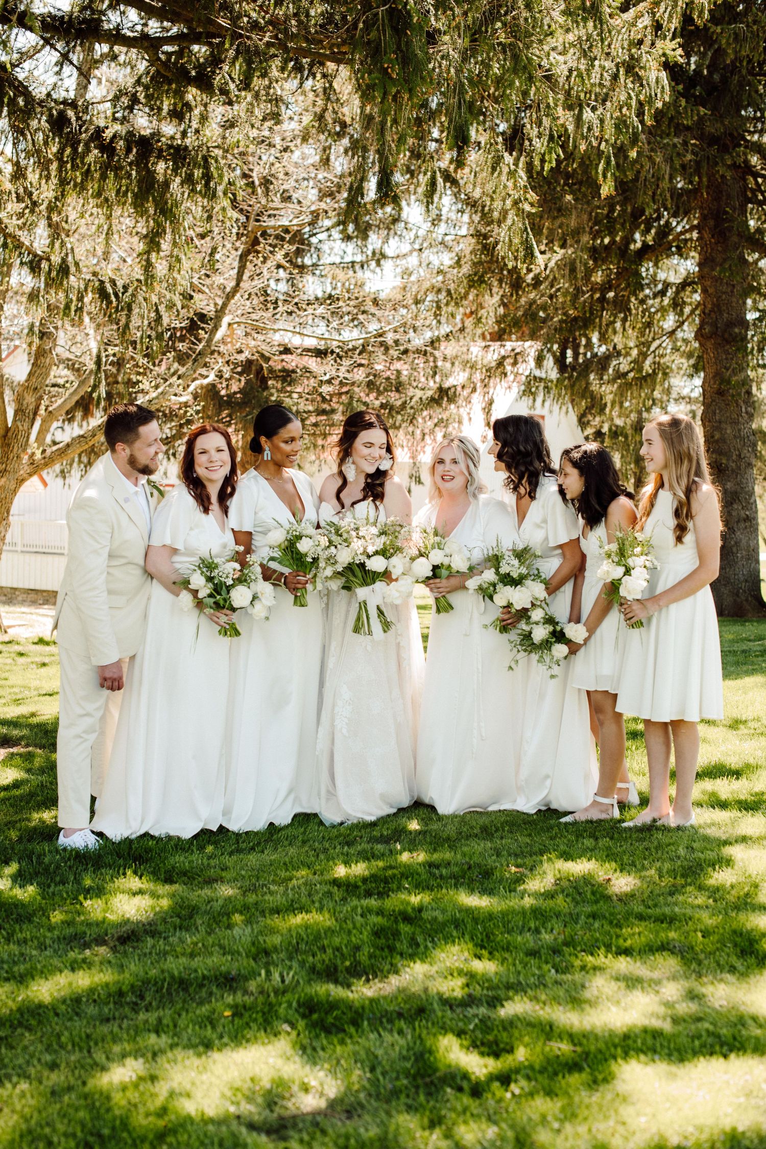 Wedding party in all-white attire posing together under evergreen trees.