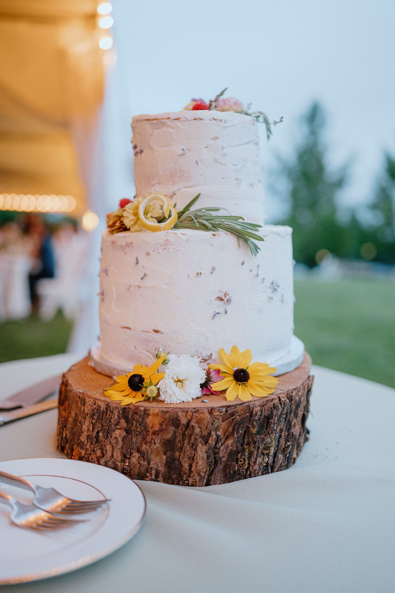 Rustic wedding cake with fresh flowers on wood slice stand at outdoor reception.