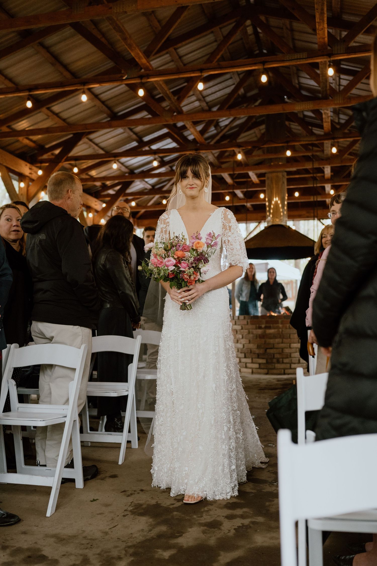 A bride in a lace wedding gown holds a colorful bouquet while walking down the aisle in a rustic venue.