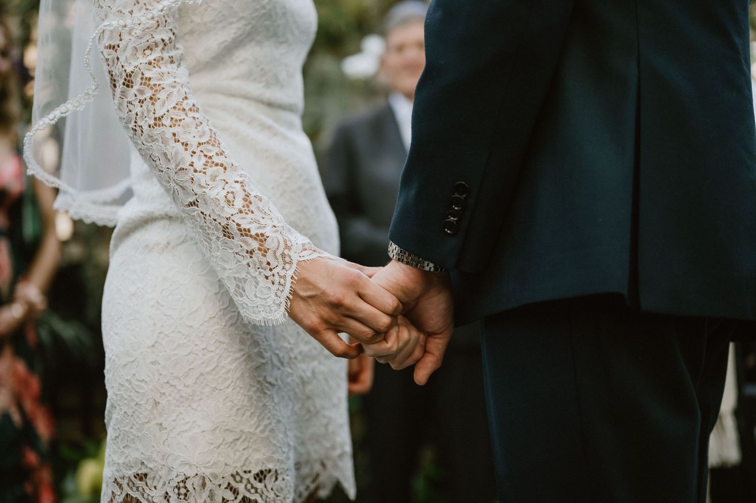 Close-up of couple holding hands during wedding ceremony in a lace dress and dark suit.