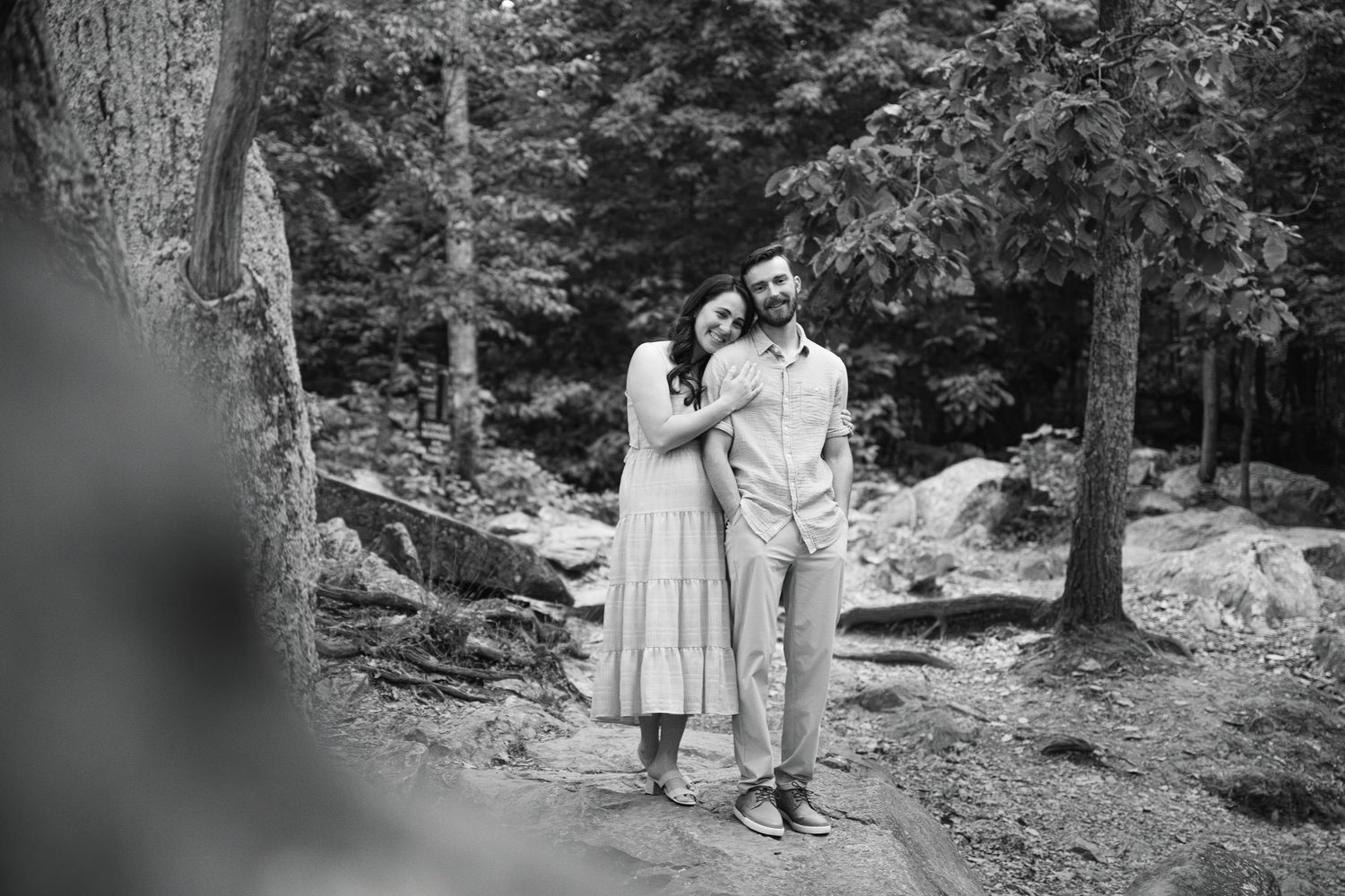 Black and white photo of a couple embracing in a forest setting with tall trees and rocks in the background.