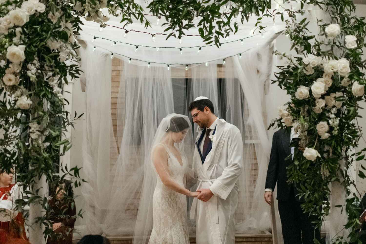 Intimate wedding ceremony moment under greenery and floral arch with white drapes and string lights.