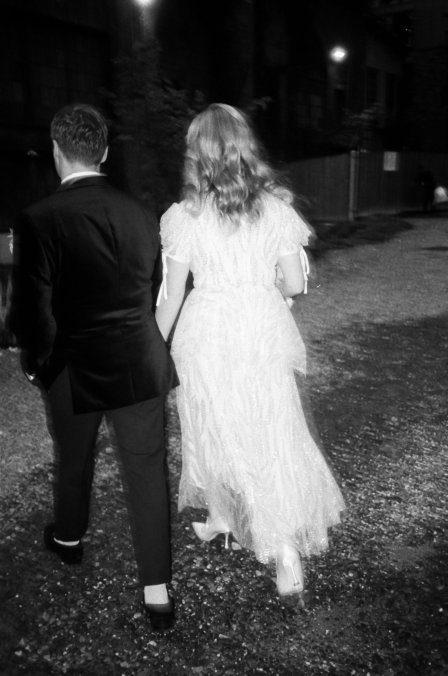 A couple walking outdoors at night, with the bride wearing a flowing white dress and groom in black suit.