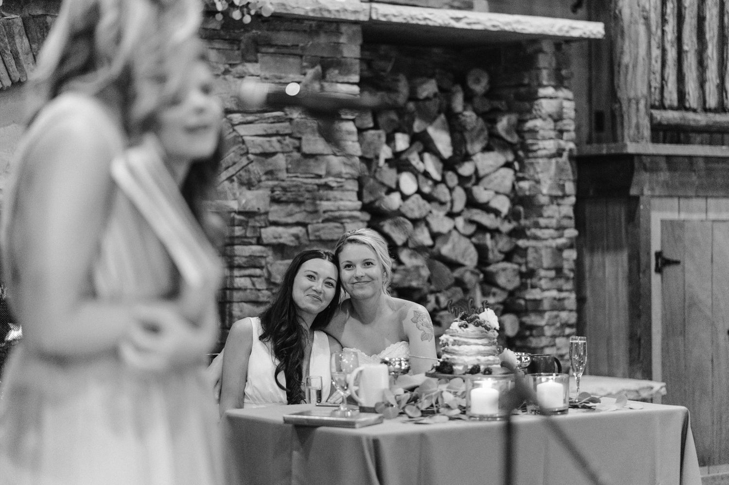 Black and white photo of wedding guests enjoying themselves at a rustic reception with stone fireplace.