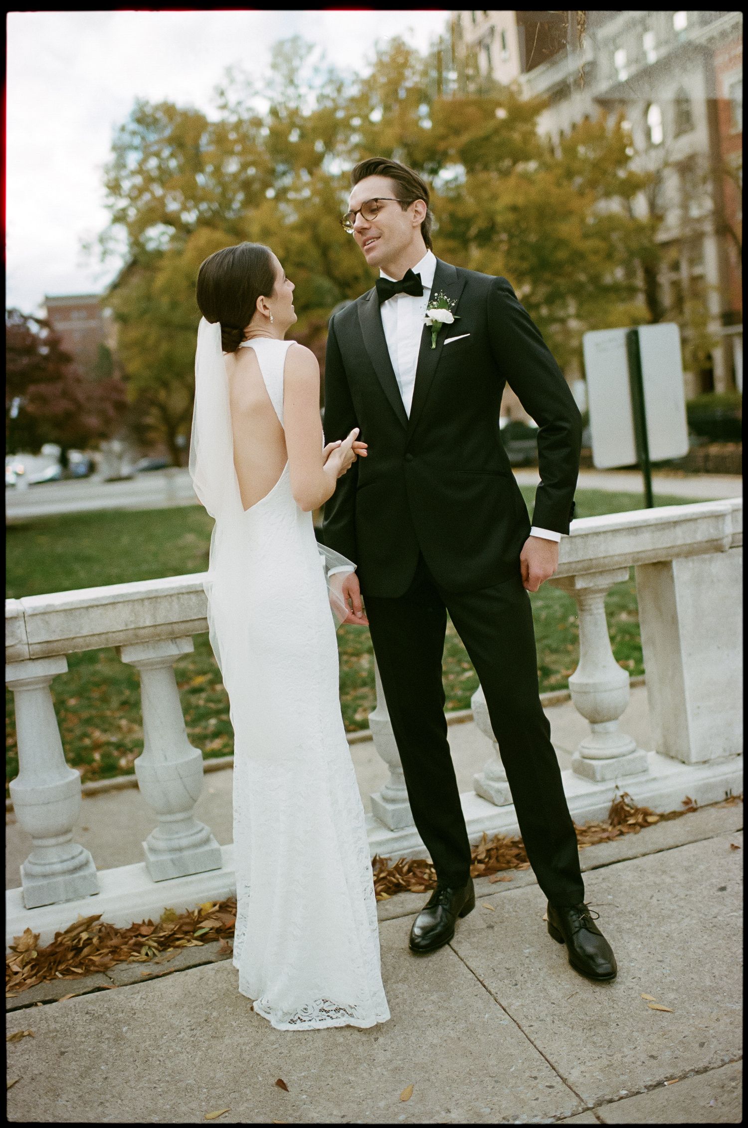 Couple in wedding attire poses on stone balcony during autumn.