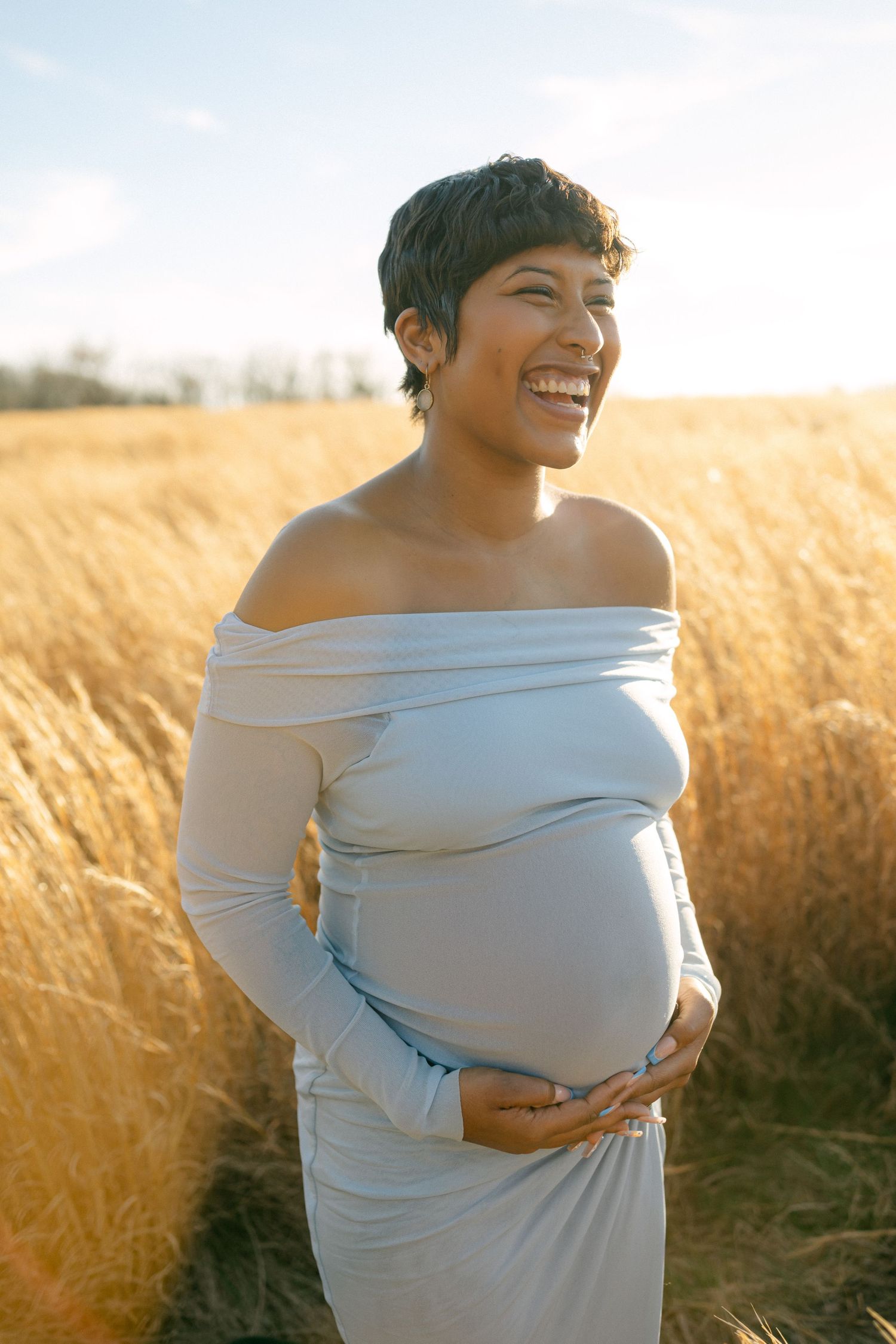 maternity photo of woman laughting holding baby bump in field with sun behind