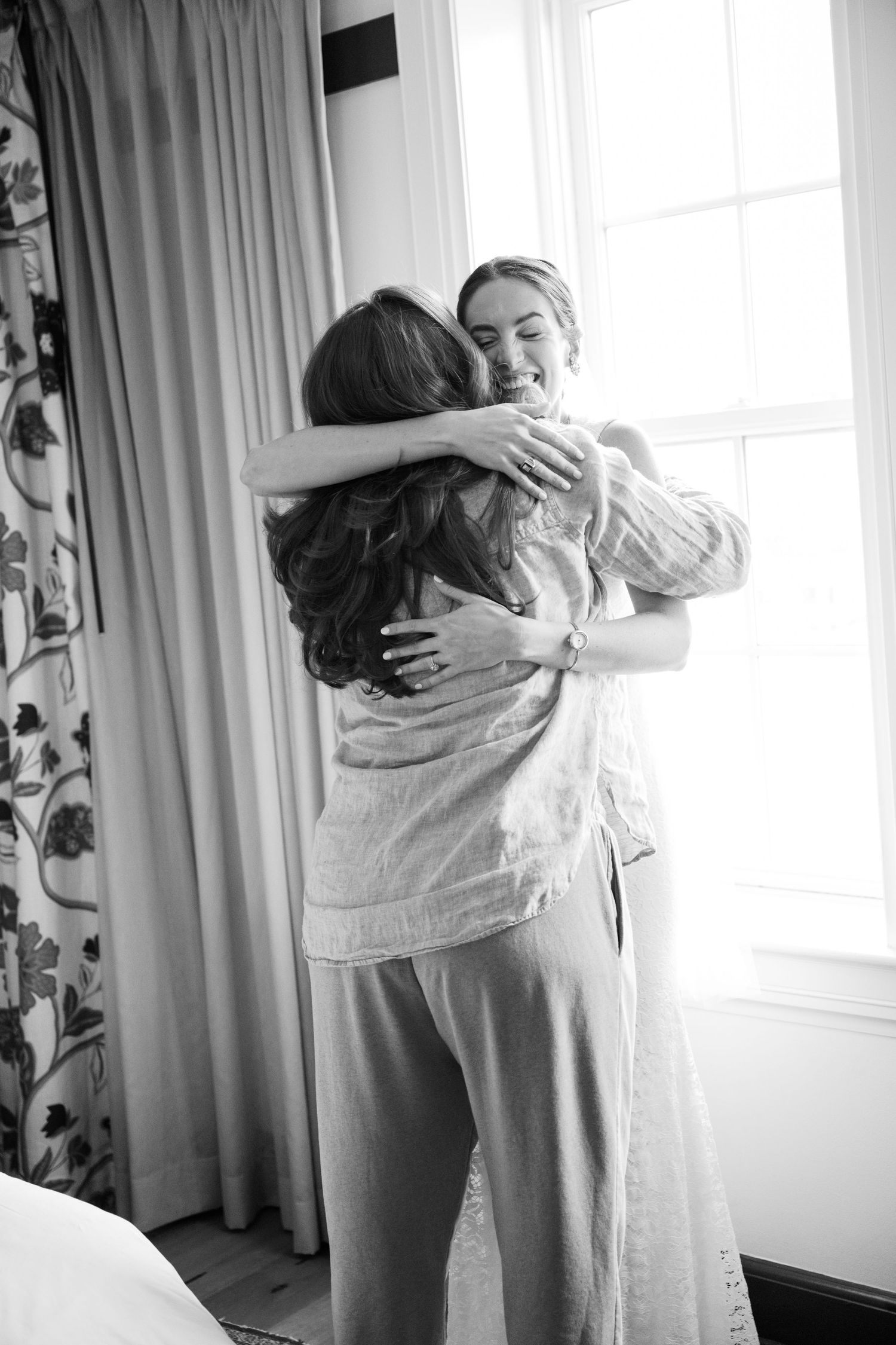 Two people sharing an emotional embrace in a bright room with floral curtains and natural window light.