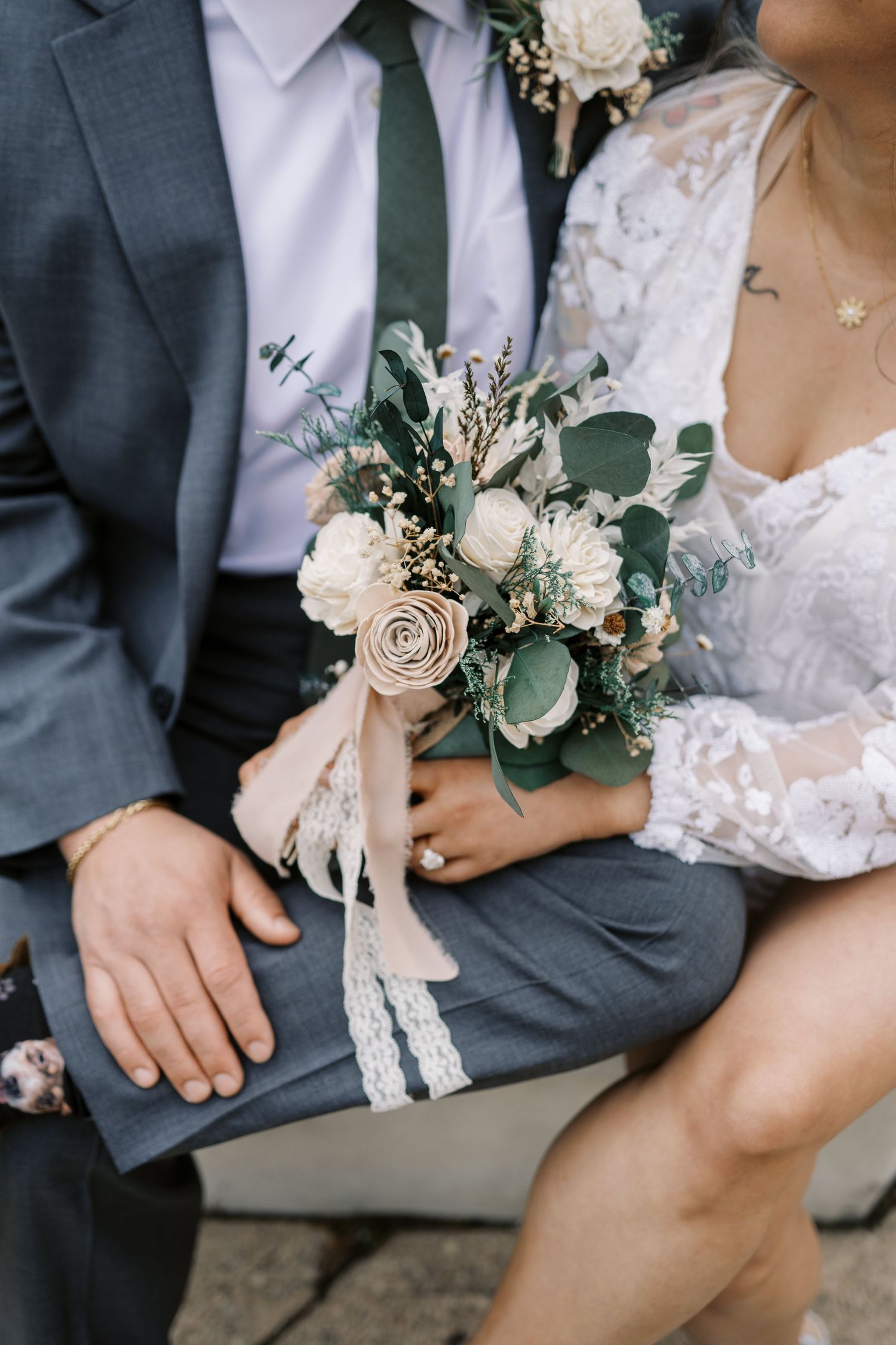A wedding bouquet with white roses and eucalyptus is held between a gray suit and lace dress.