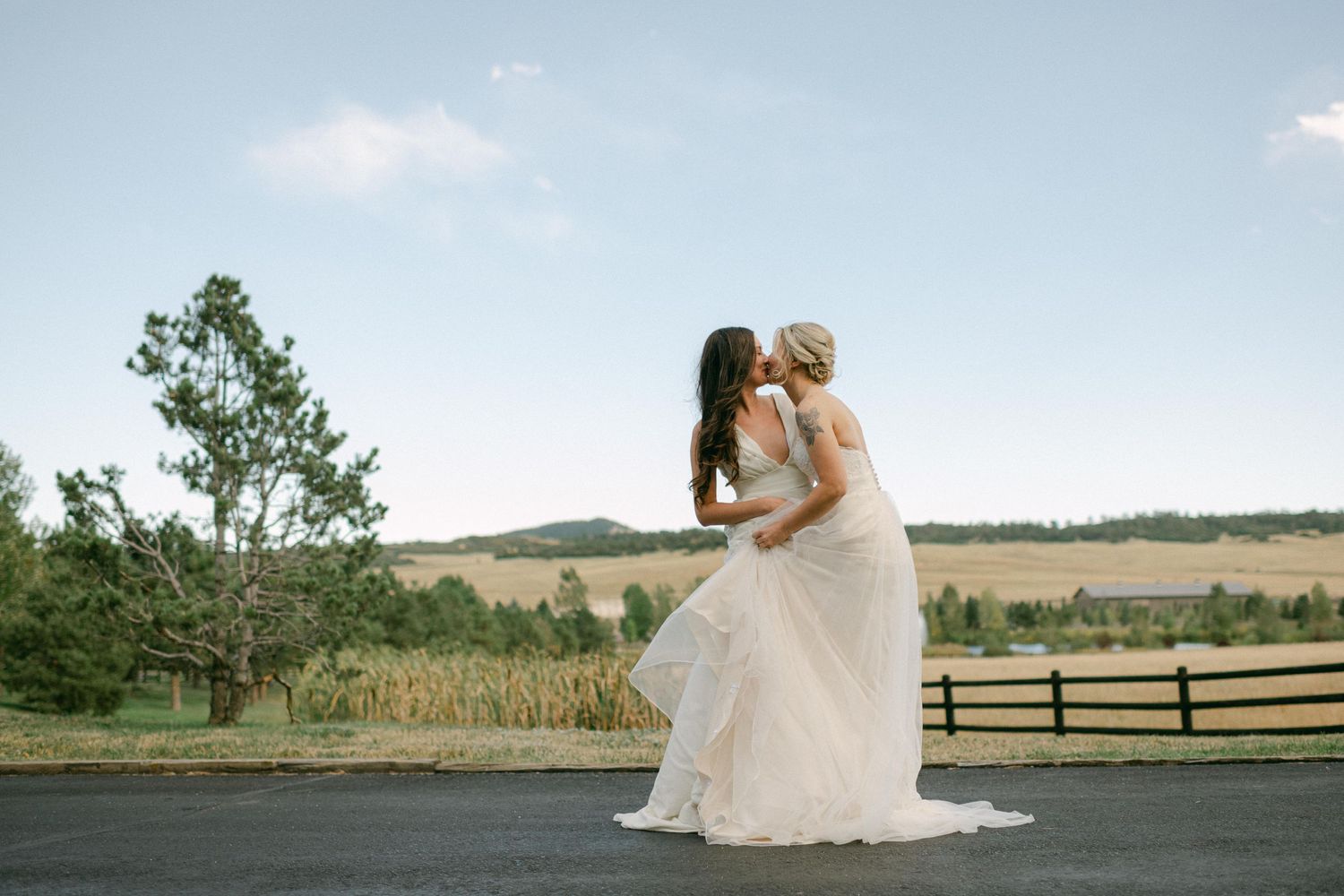 Two people in white wedding dresses embrace on a road with scenic countryside views.