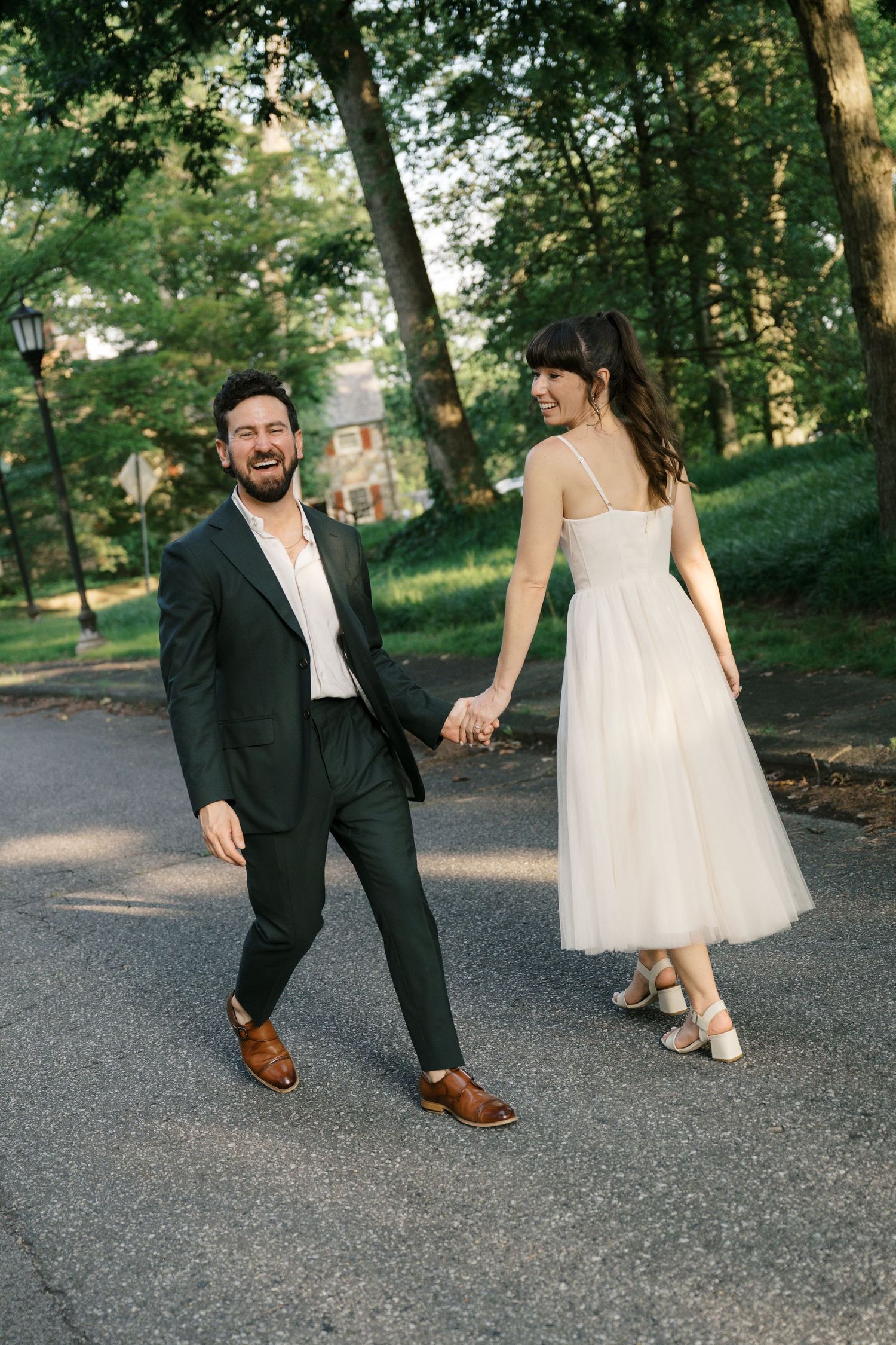 A couple in wedding attire shares playful moments on a paved path surrounded by lush green trees.