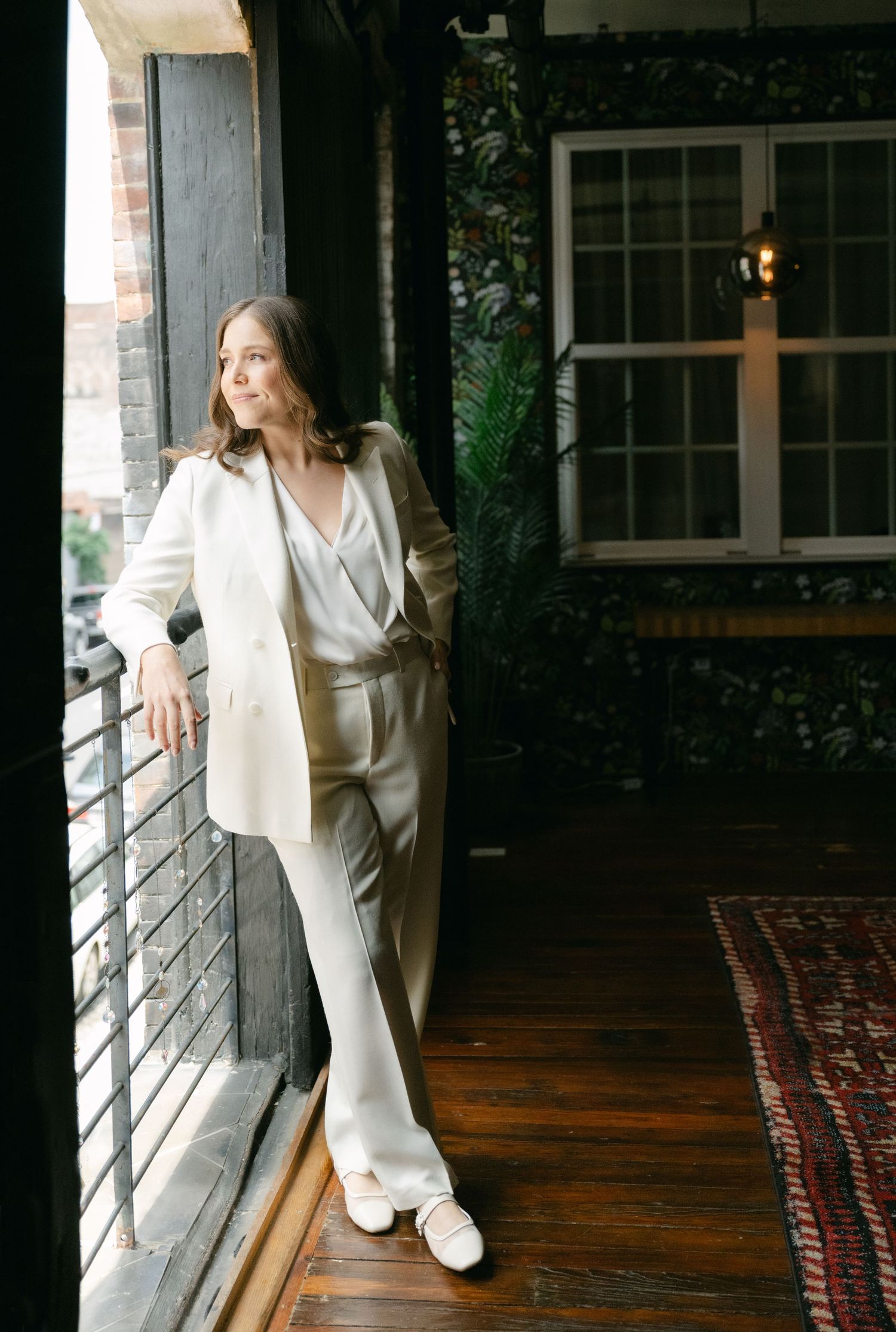 Figure in elegant white pantsuit poses by sunny window in artistic portrait series.