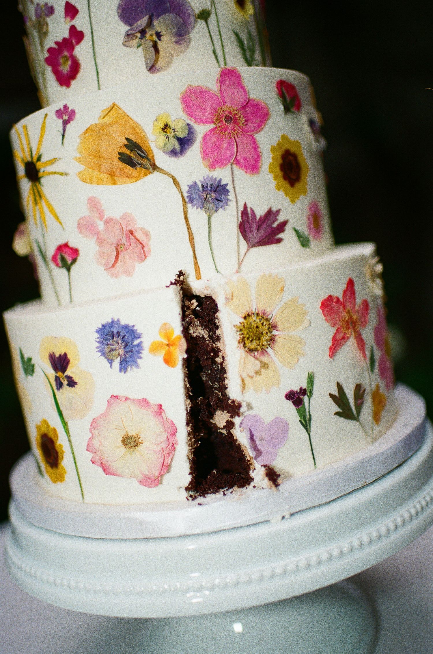 A white wedding cake decorated with pressed edible flowers in pink, purple, and yellow on display.