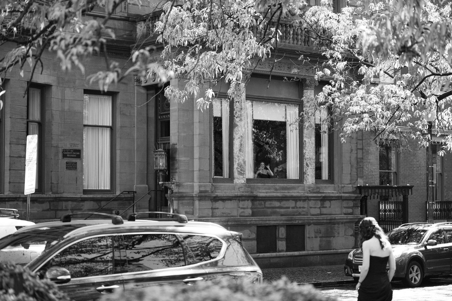 Black and white urban street scene showing brownstone building facade with parked cars and tree branches overhead.