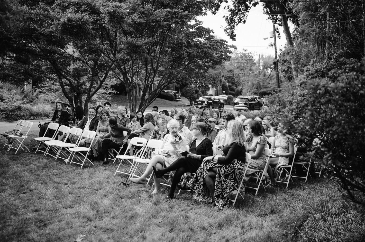 Black and white photo of wedding guests seated in rows of white chairs during an outdoor ceremony.