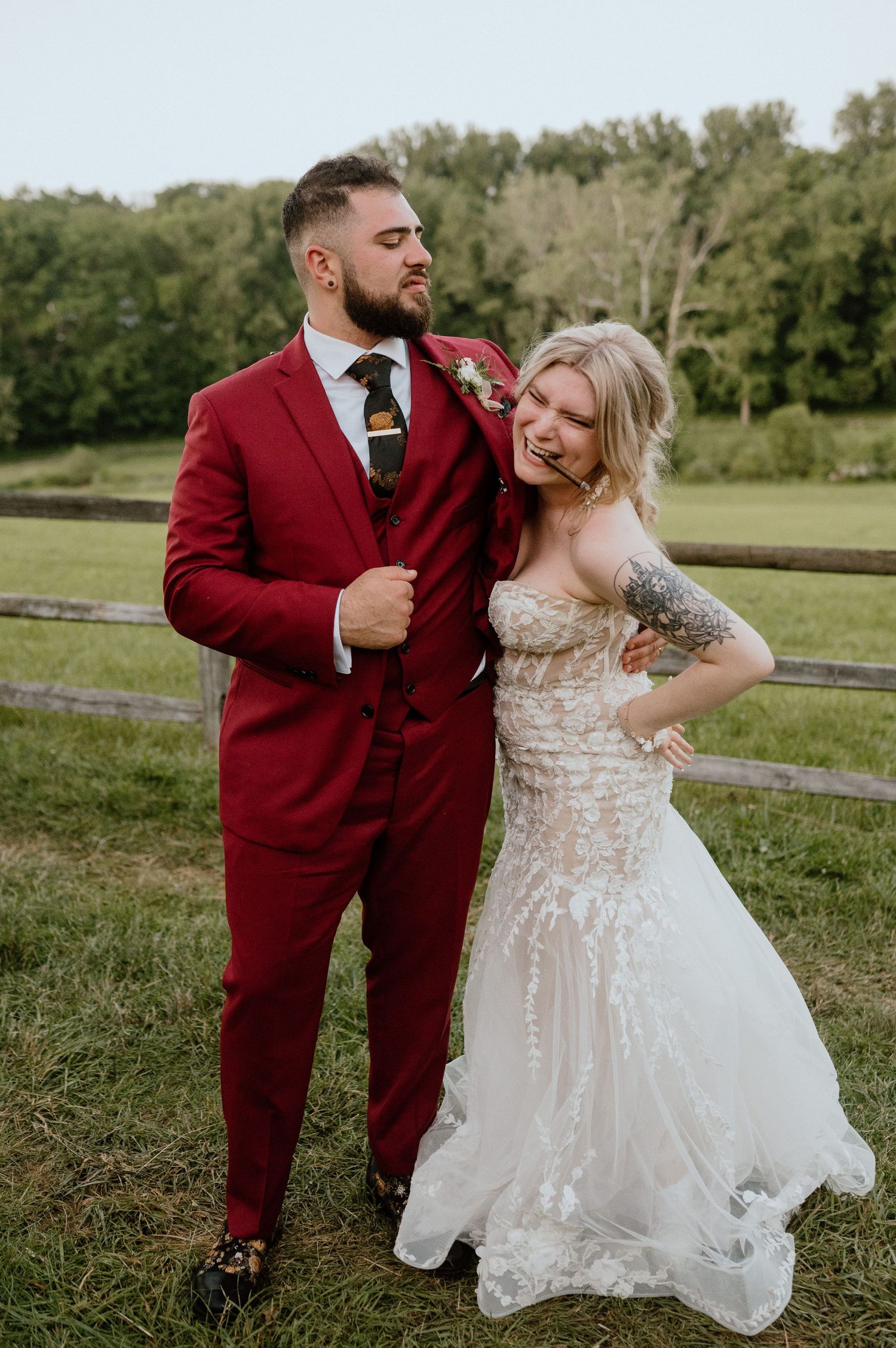 A couple in wedding attire poses by a wooden fence in a countryside setting.