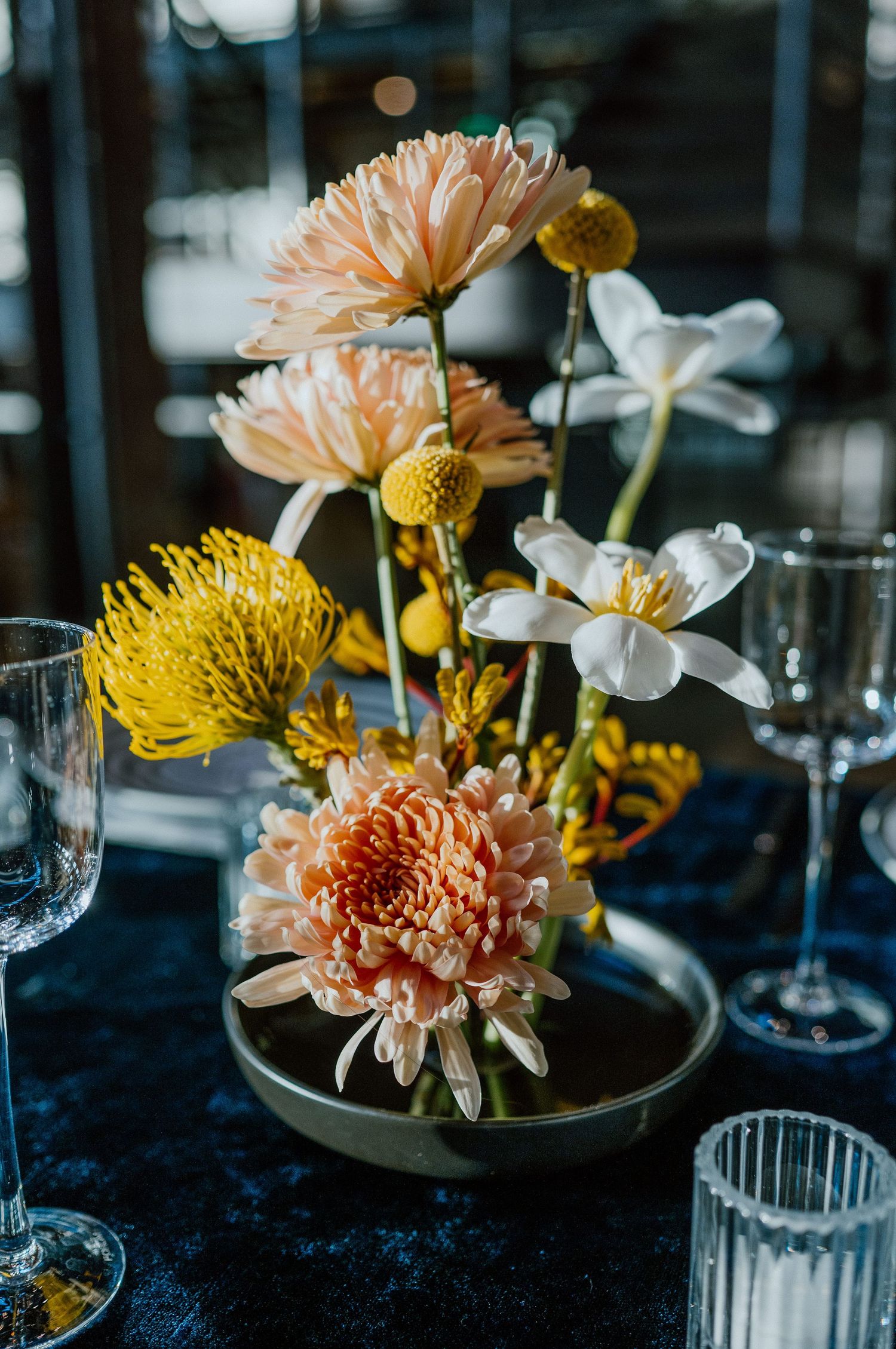 Elegant floral arrangement with peach dahlias, yellow pincushion flowers and white blooms in a modern vase setting.