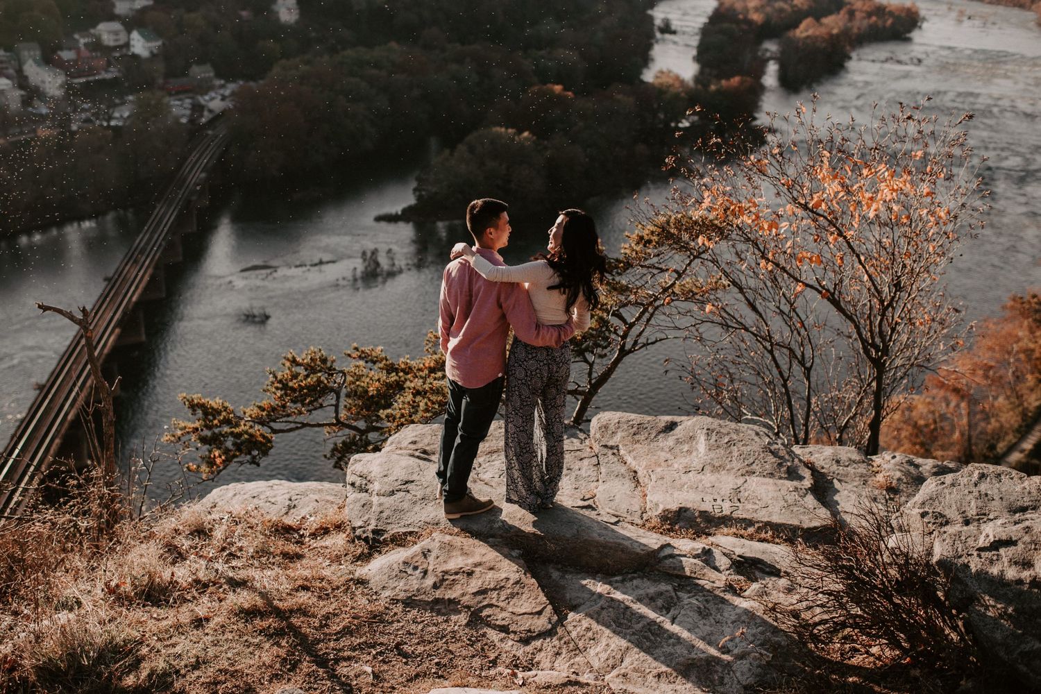 Couple standing on rocky cliff edge with scenic river and autumn foliage view.