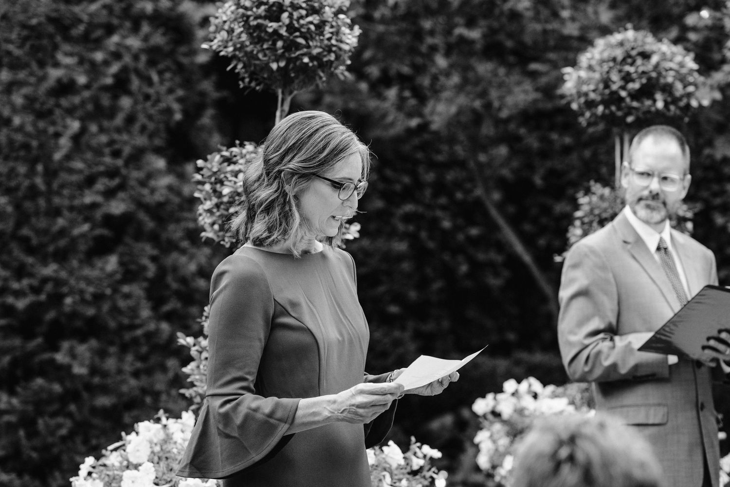 A wedding reading takes place during an outdoor ceremony surrounded by greenery and flowers.