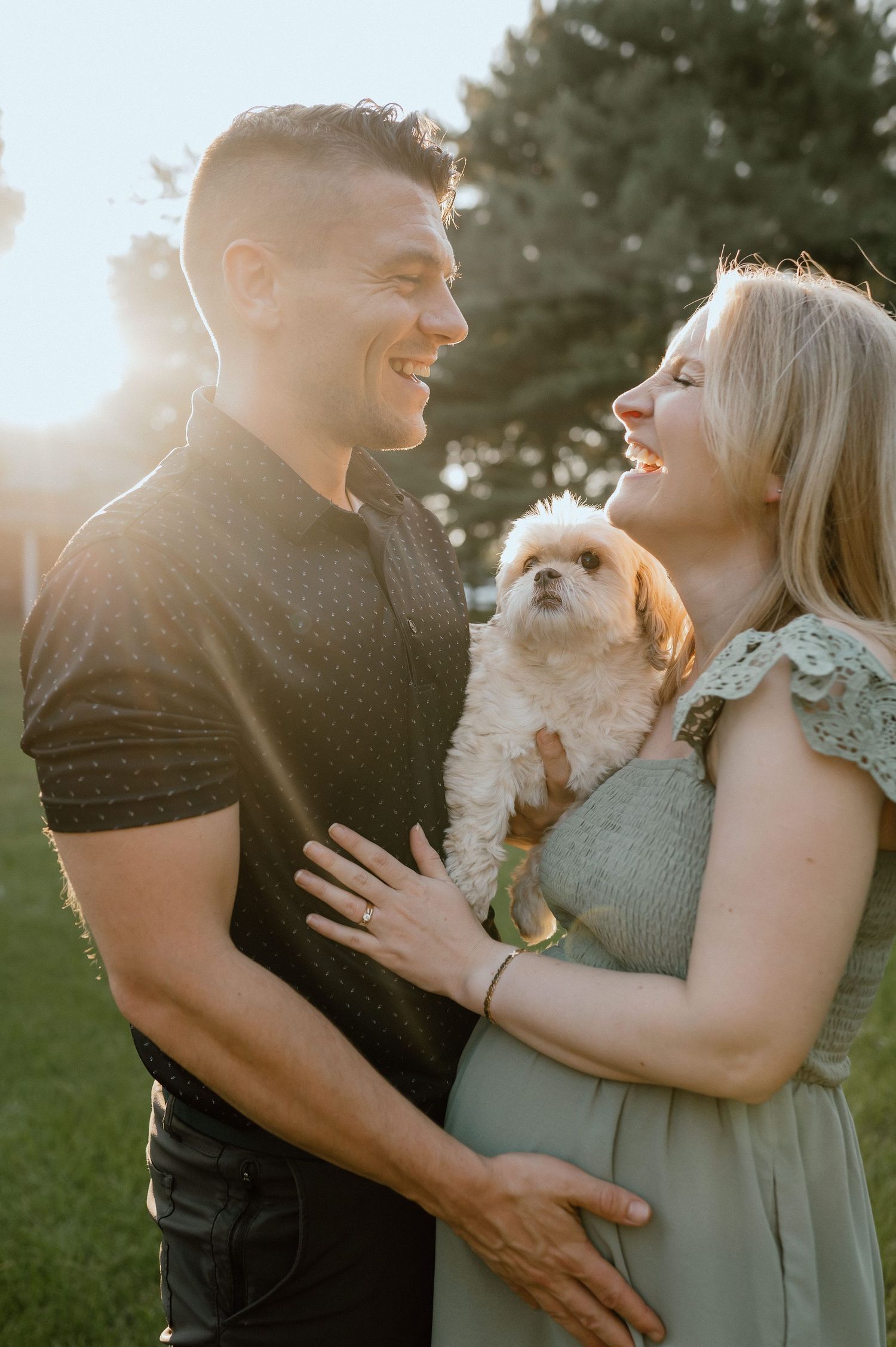 Couple holding small white dog while sharing a joyful moment in evening sunlight.