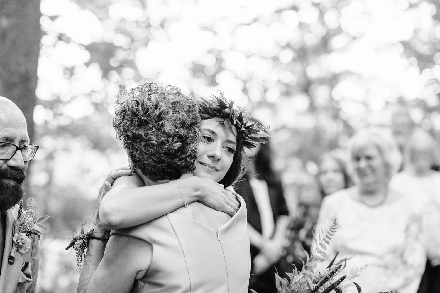 Black and white photograph captures an emotional embrace during a forest wedding celebration.