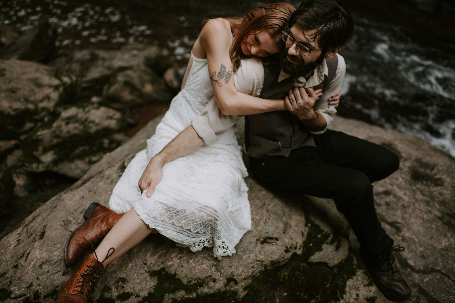 A couple in wedding attire sits intimately on a rocky surface in nature.
