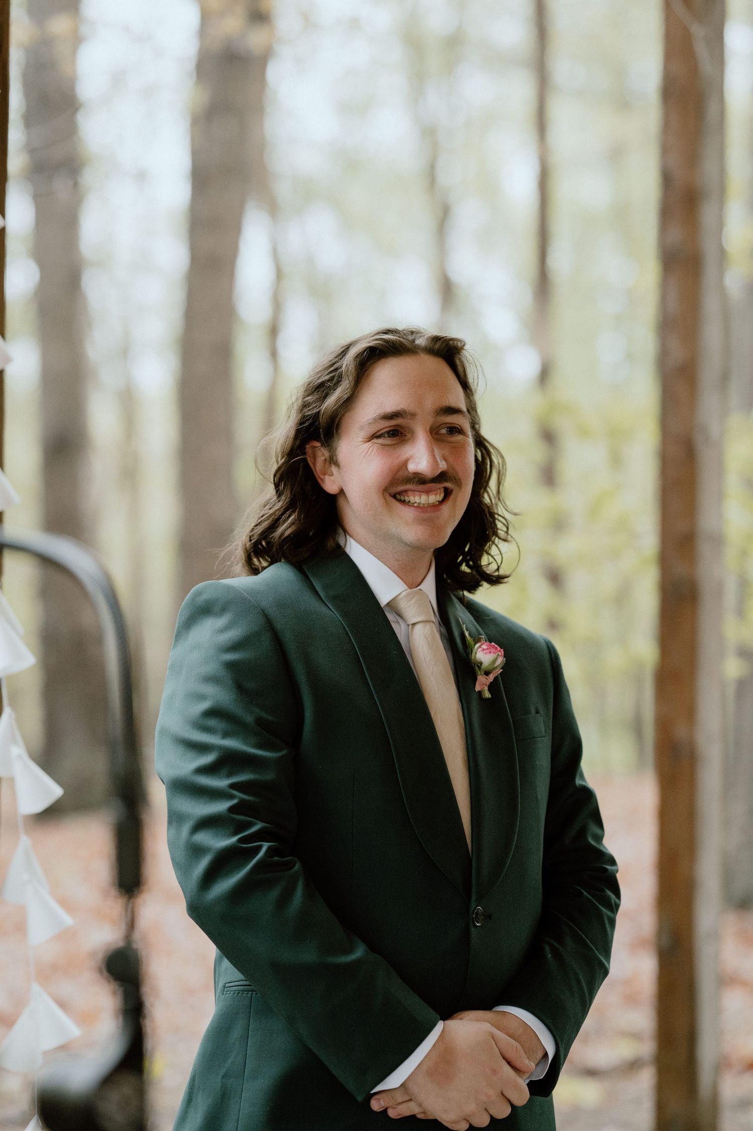 A groomsman with long wavy hair and a mustache smiles while wearing a dark green suit jacket and cream tie.